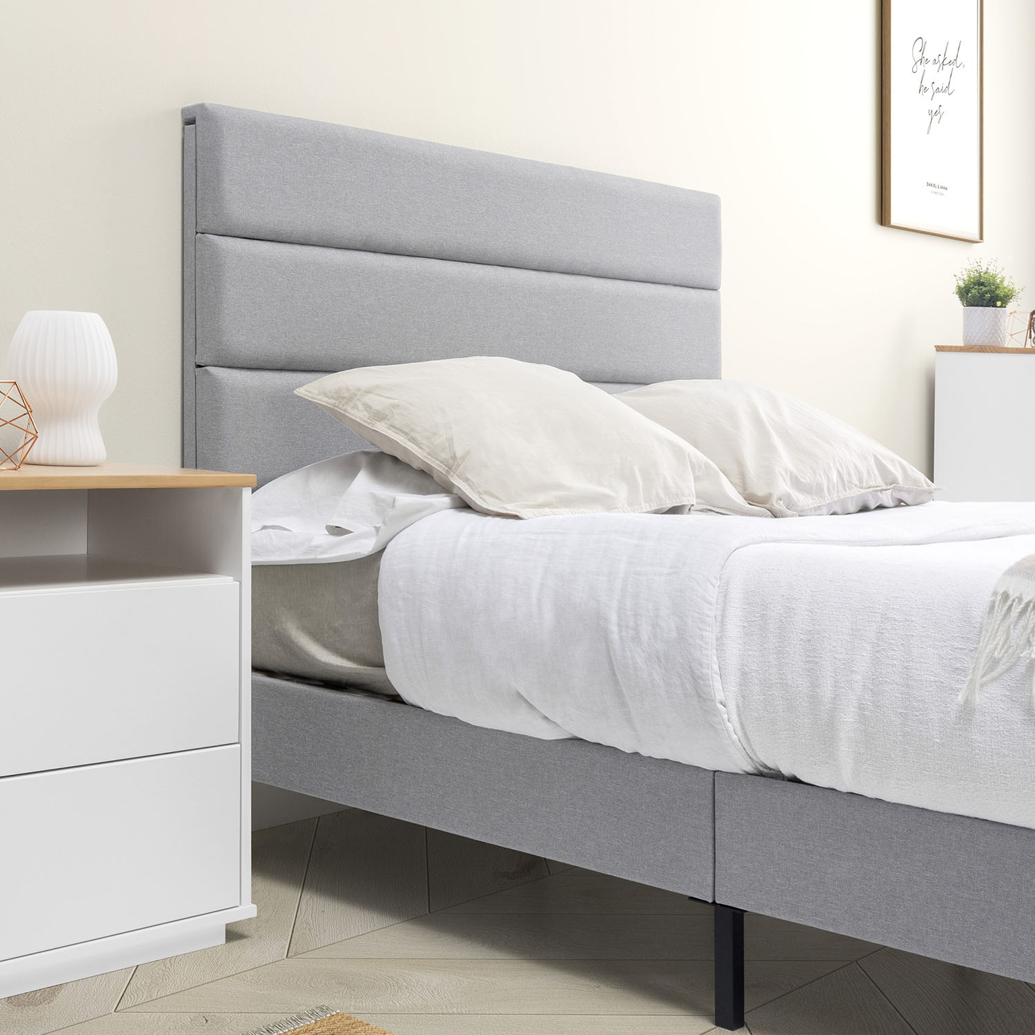 Una cama tapizada gris con sábanas y almohadas blancas se encuentra junto a una mesita de noche blanca.