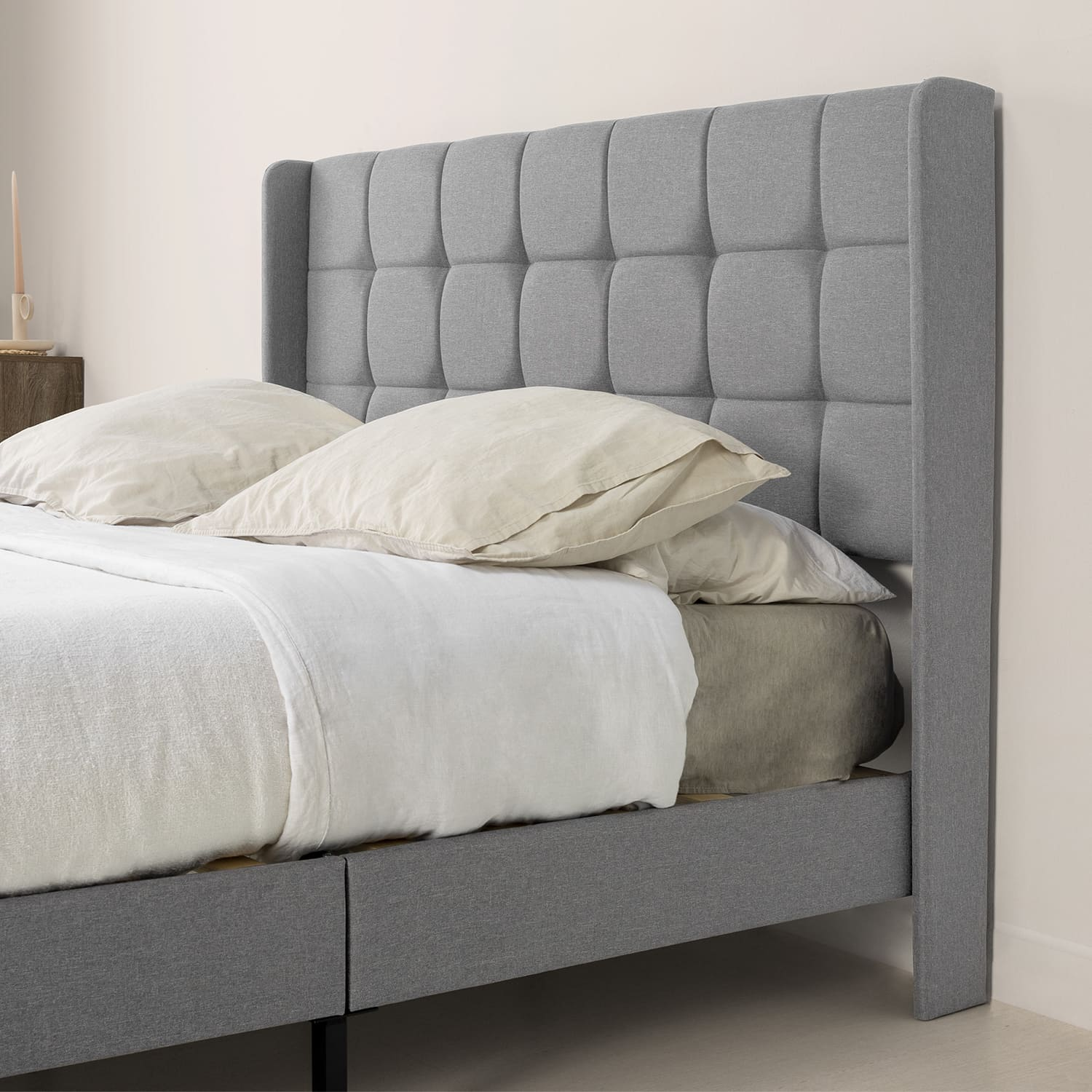 Una cama tapizada gris con cabecero y barandillas laterales, adornada con ropa de cama blanca y beige.
