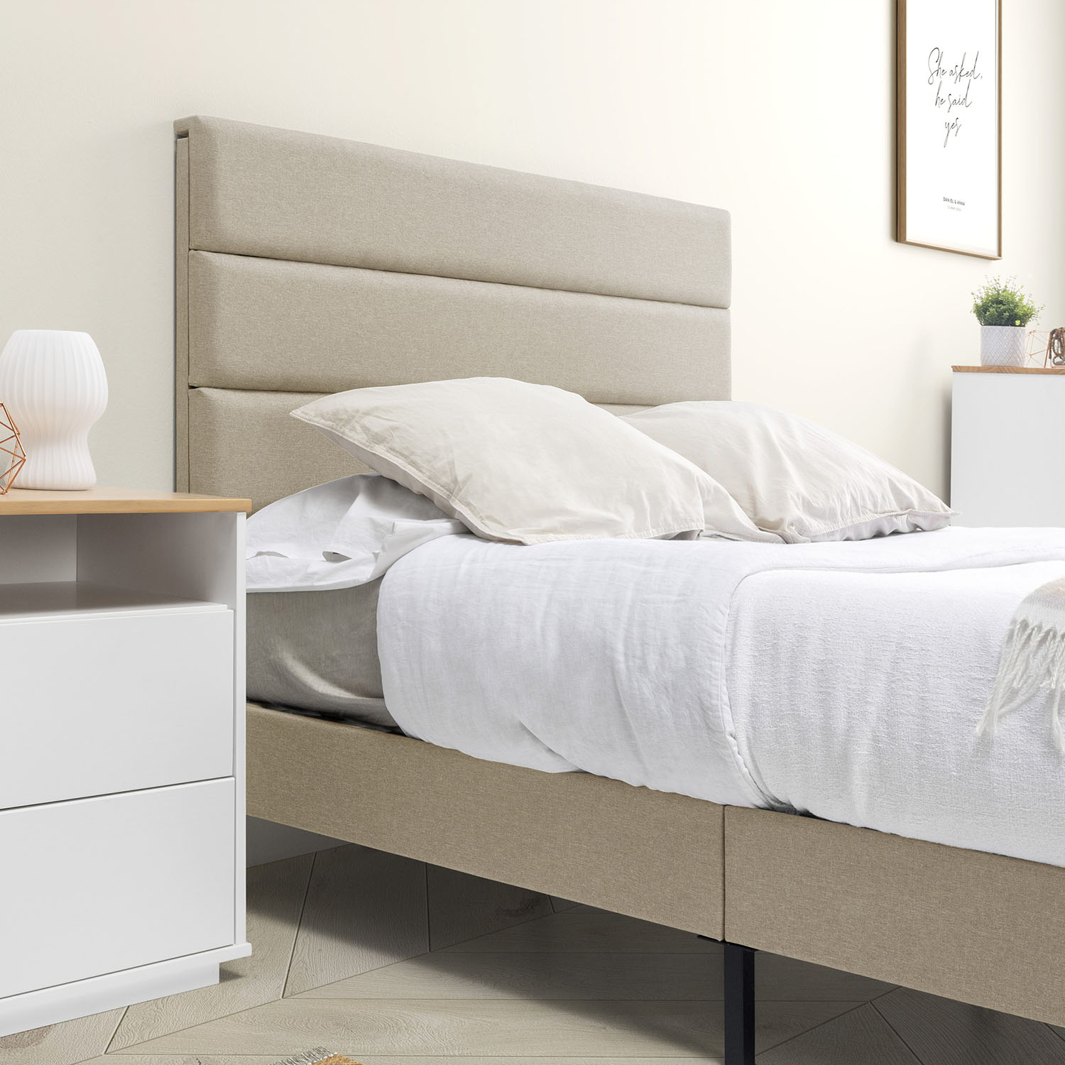 Una cama con cabecero tapizado beige y mesita de noche al lado.