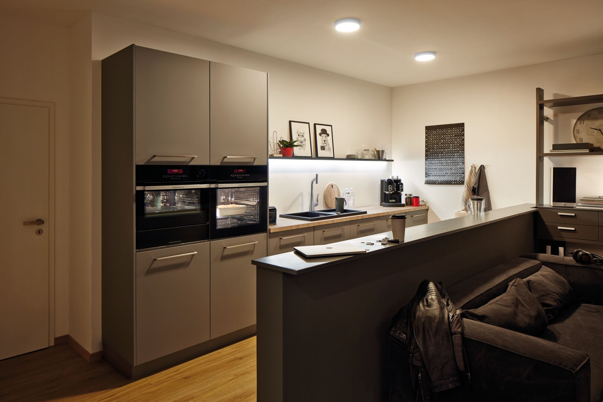 Cucina moderna con doppio forno, lavello e macchina da caffè. Zona soggiorno con divano e giacca.