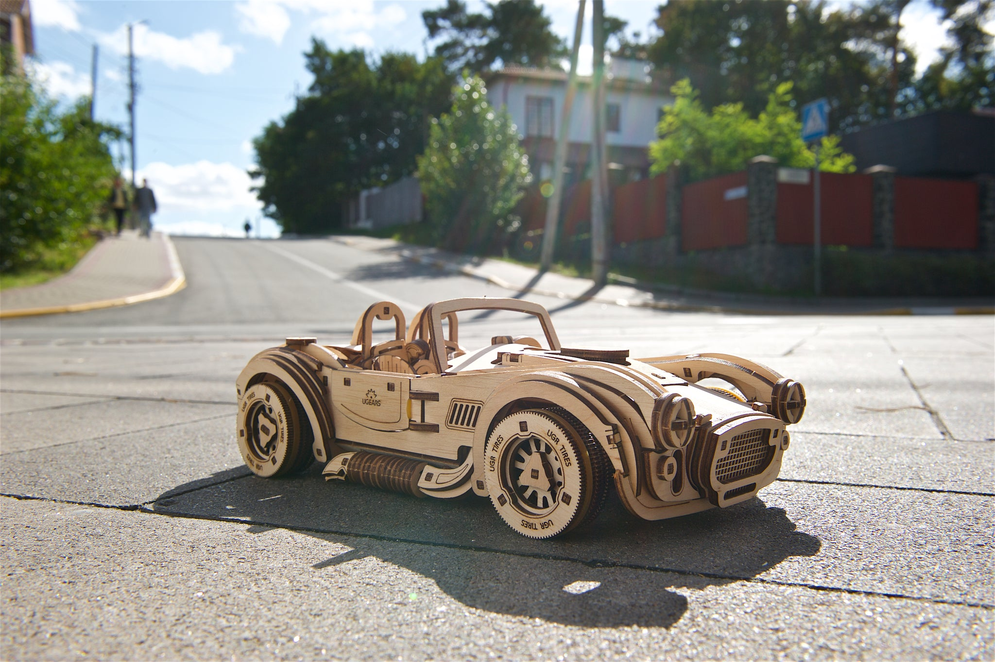 Ein hölzernes Modellauto steht auf einer Straße. Im Hintergrund sind Bäume und Häuser.