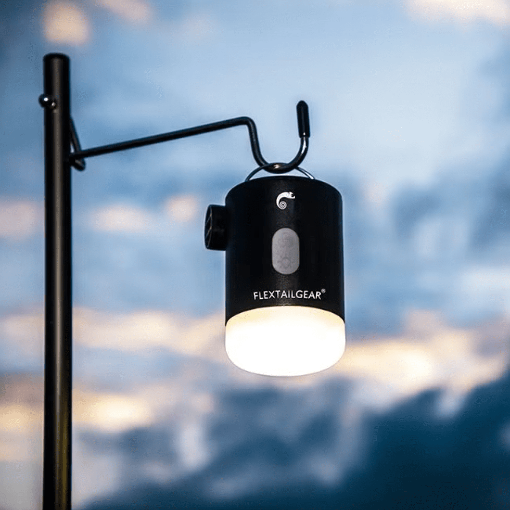 Eine schwarze Laterne mit heller Beleuchtung hängt an einem Gestell vor einem bewölkten Himmel.