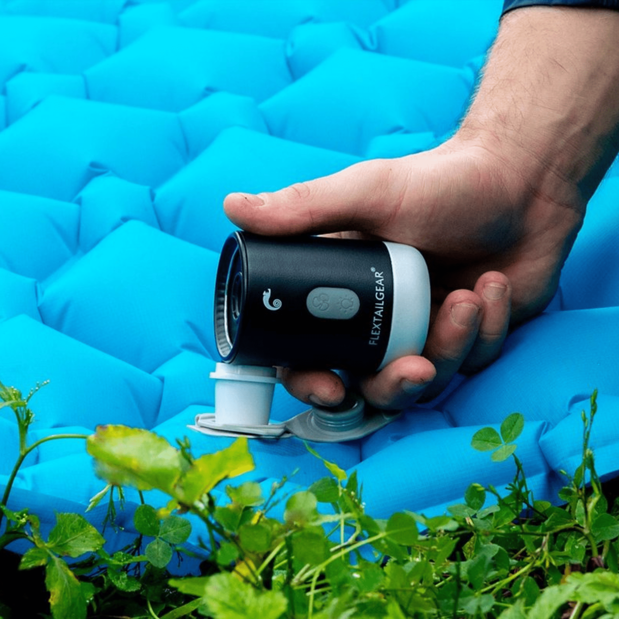 Eine Hand hält eine Pumpe auf einer blauen Isomatte, umgeben von grünem Gras.