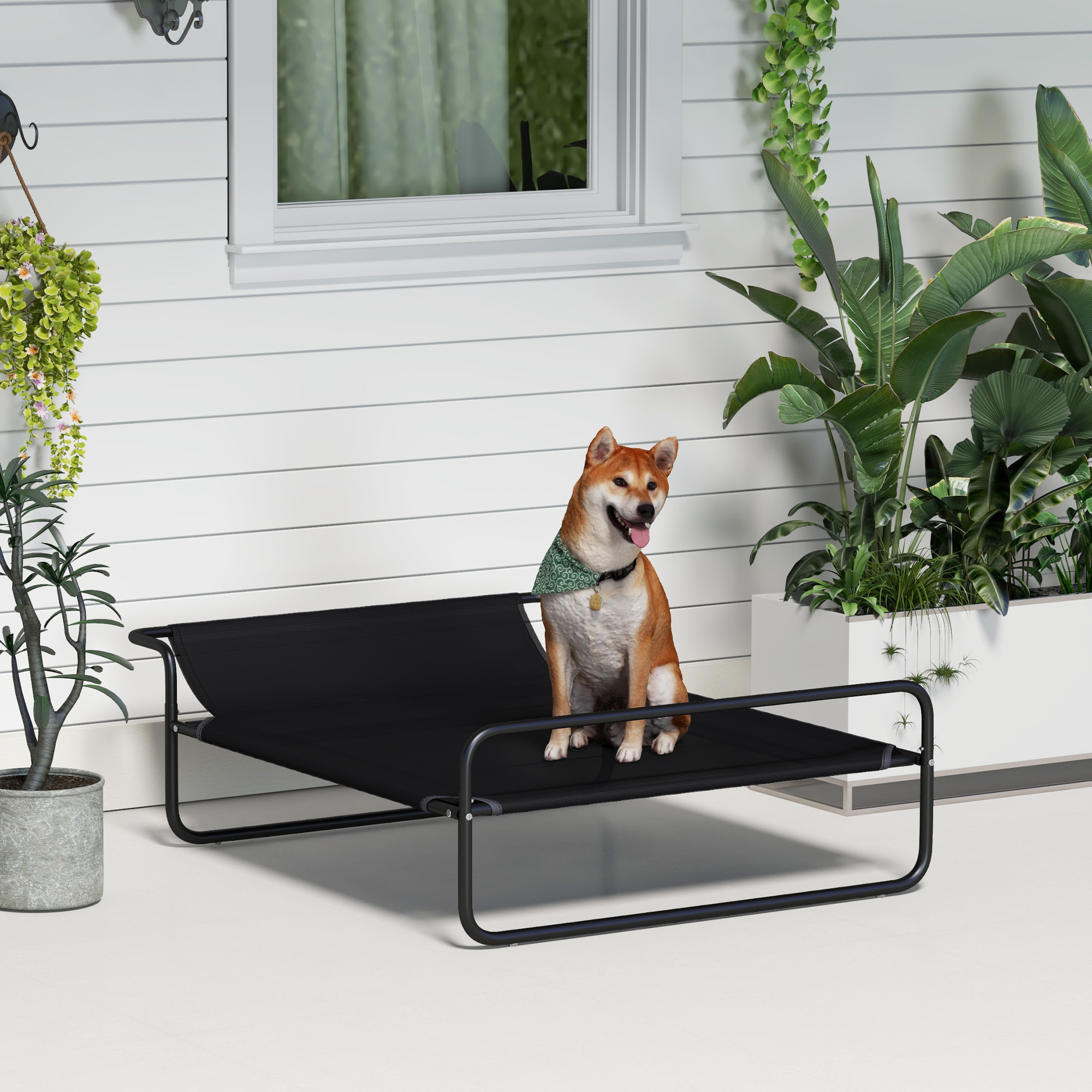 Un perro Shiba Inu se sienta en una cama de perro negra al aire libre junto a plantas.