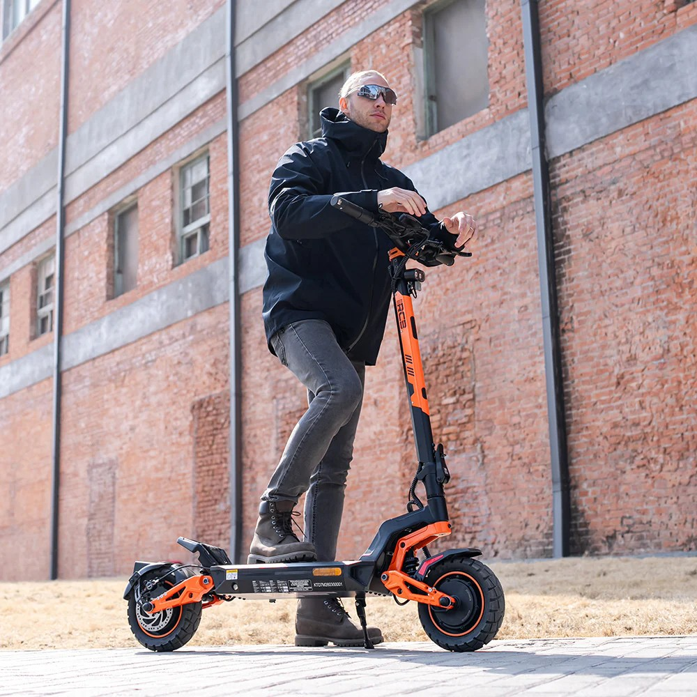 Ein Mann in dunkler Kleidung fährt einen orange-schwarzen Elektroroller vor einer Backsteinwand.