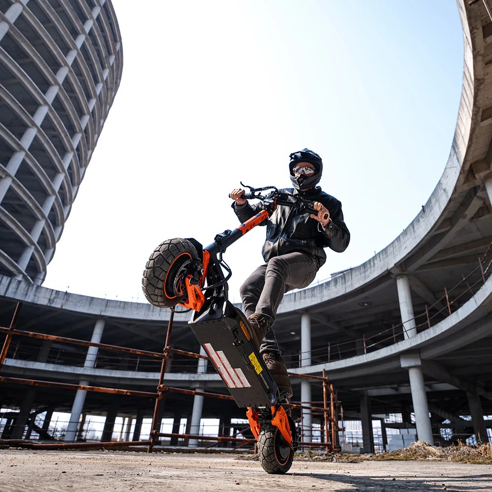 Ein Mann auf einem orangefarbenen Elektroroller macht einen Wheelie vor einer Betonstruktur.