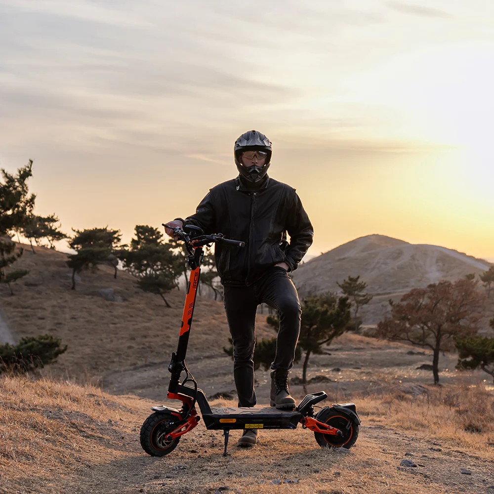 Ein Mann in Lederjacke und Helm steht neben einem schwarzen und orangen E-Scooter im Freien.