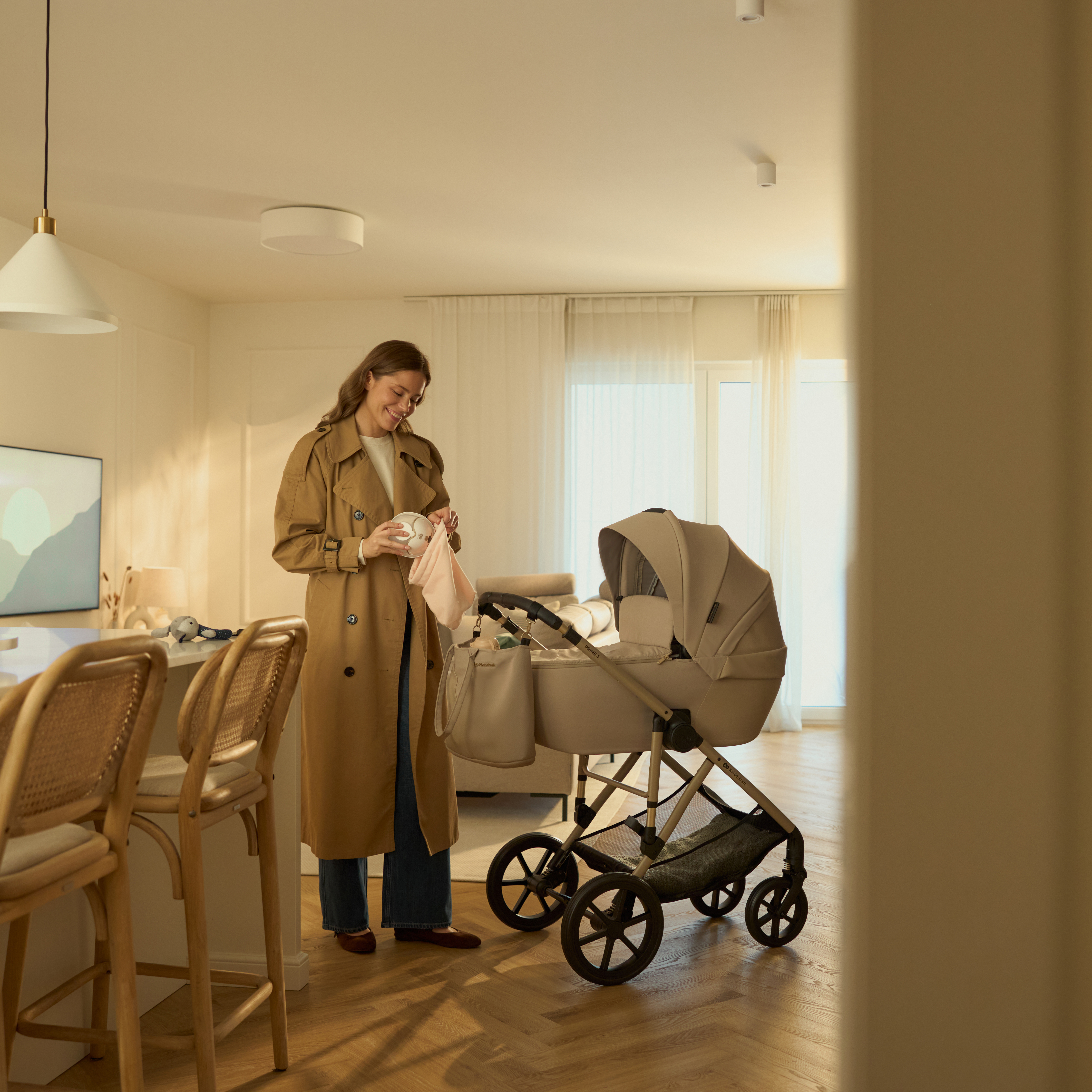 Eine Frau in einem beigen Trenchcoat steht neben einem Kinderwagen in einem hellen Raum.