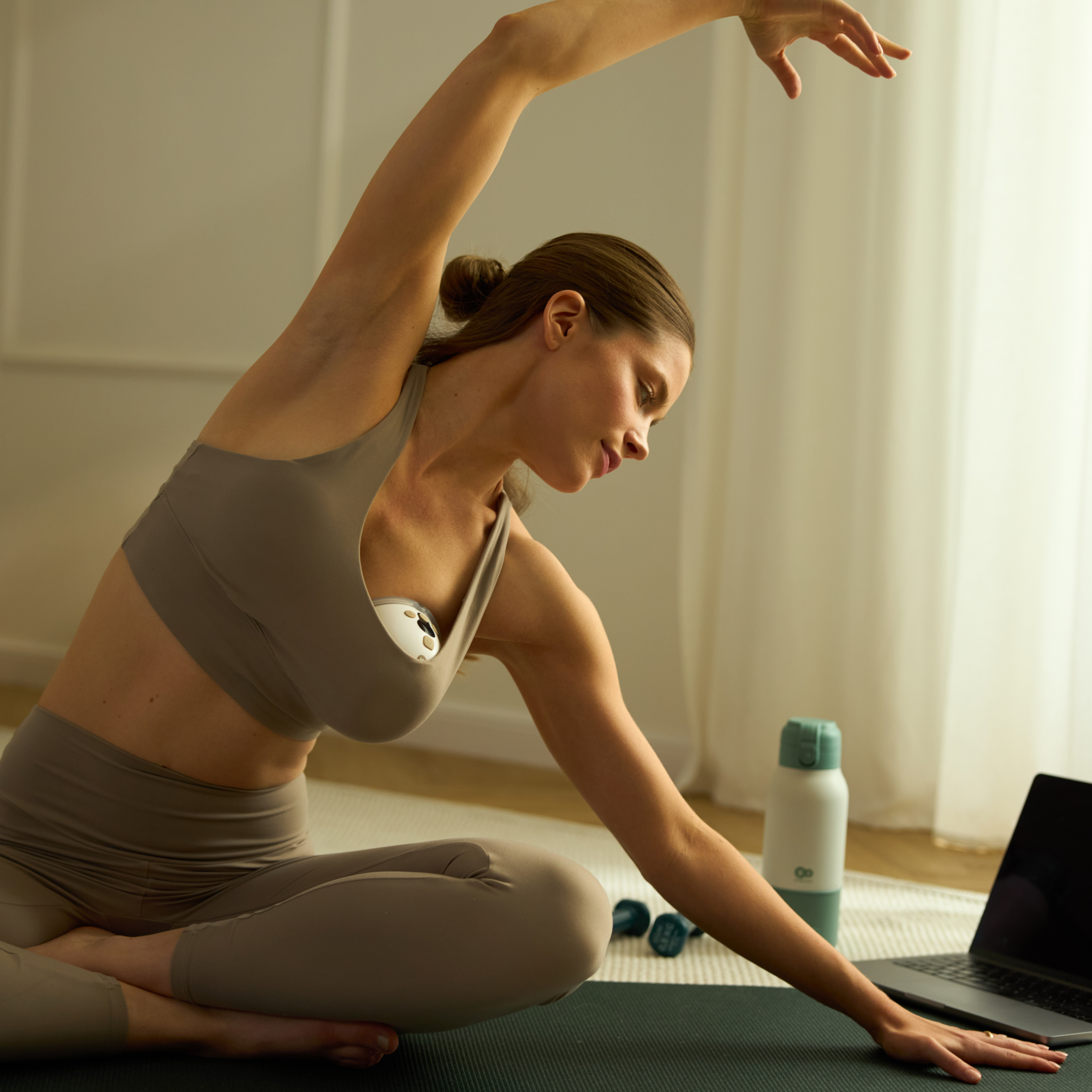 Eine Frau in Sportkleidung dehnt sich auf einer Matte, ein Laptop und eine Wasserflasche daneben.