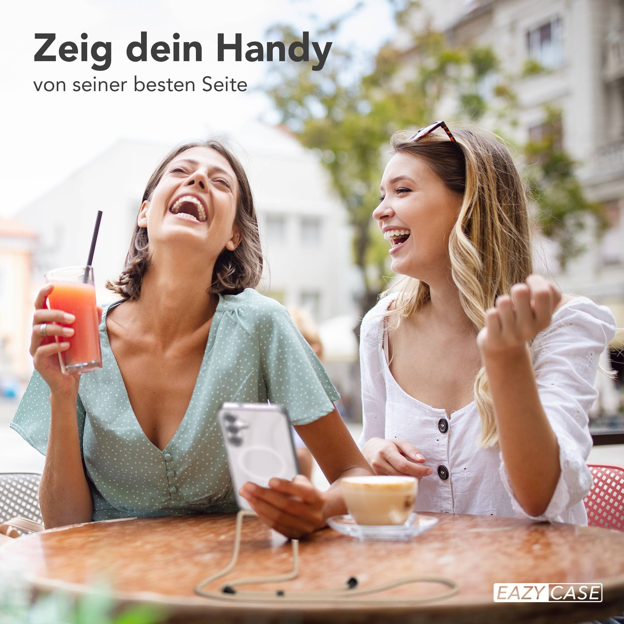 Zwei lachende Frauen sitzen im Freien an einem Tisch mit Getränken und einem Handy.