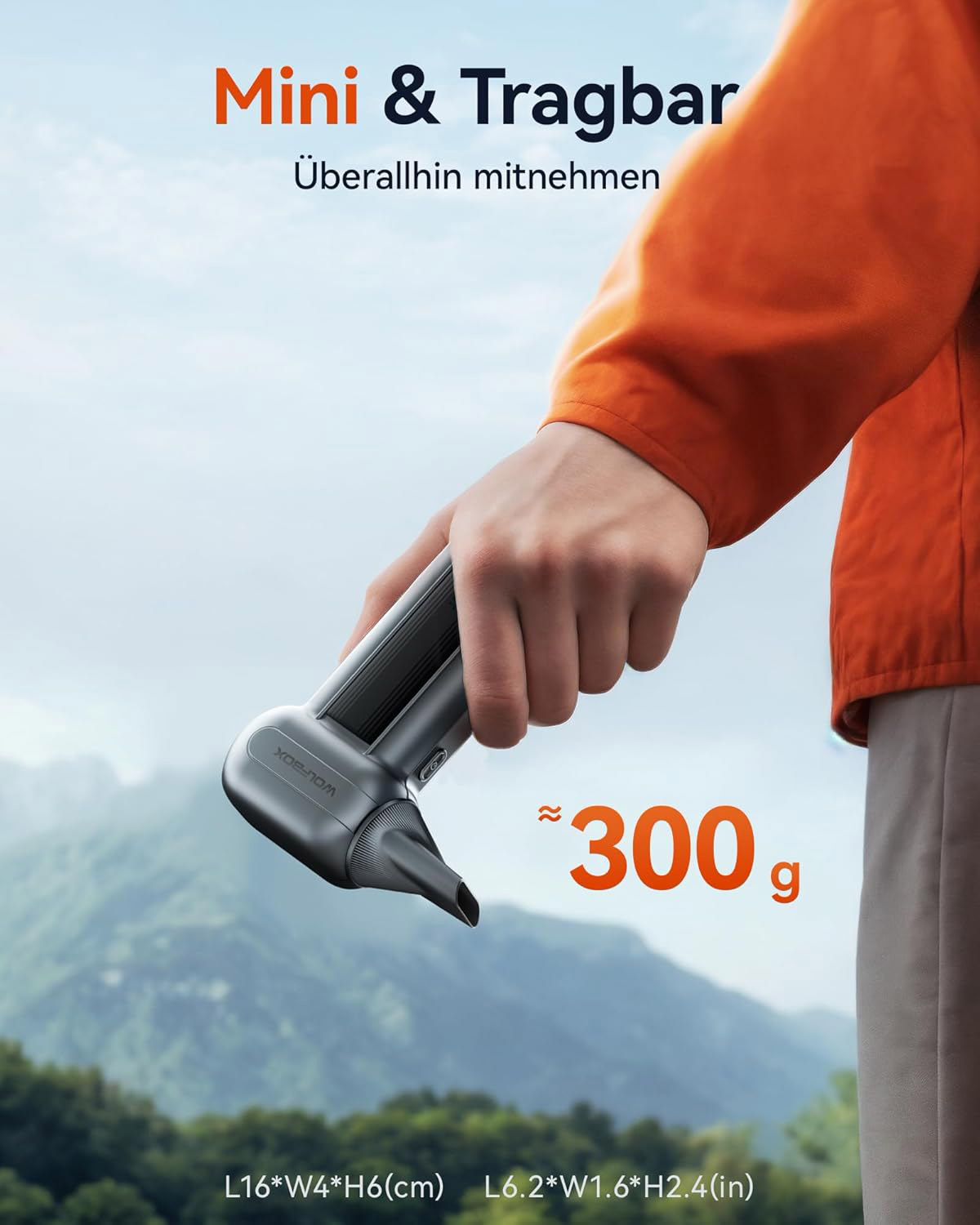 Eine Hand hält ein kleines, graues Gerät vor einer Berglandschaft. Es wiegt 300g.