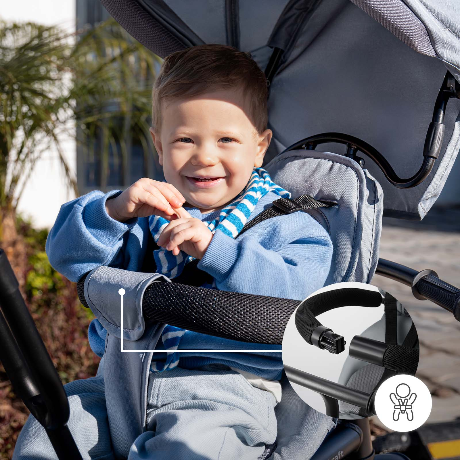 Ein lächelnder Junge sitzt in einem grauen Kinderwagen mit einem blauen gestreiften Schal.