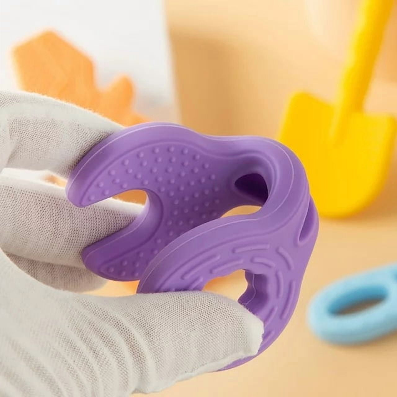 Eine Hand in einem weißen Handschuh hält ein lila Spielzeug. Gelbe und blaue Spielzeuge im Hintergrund.