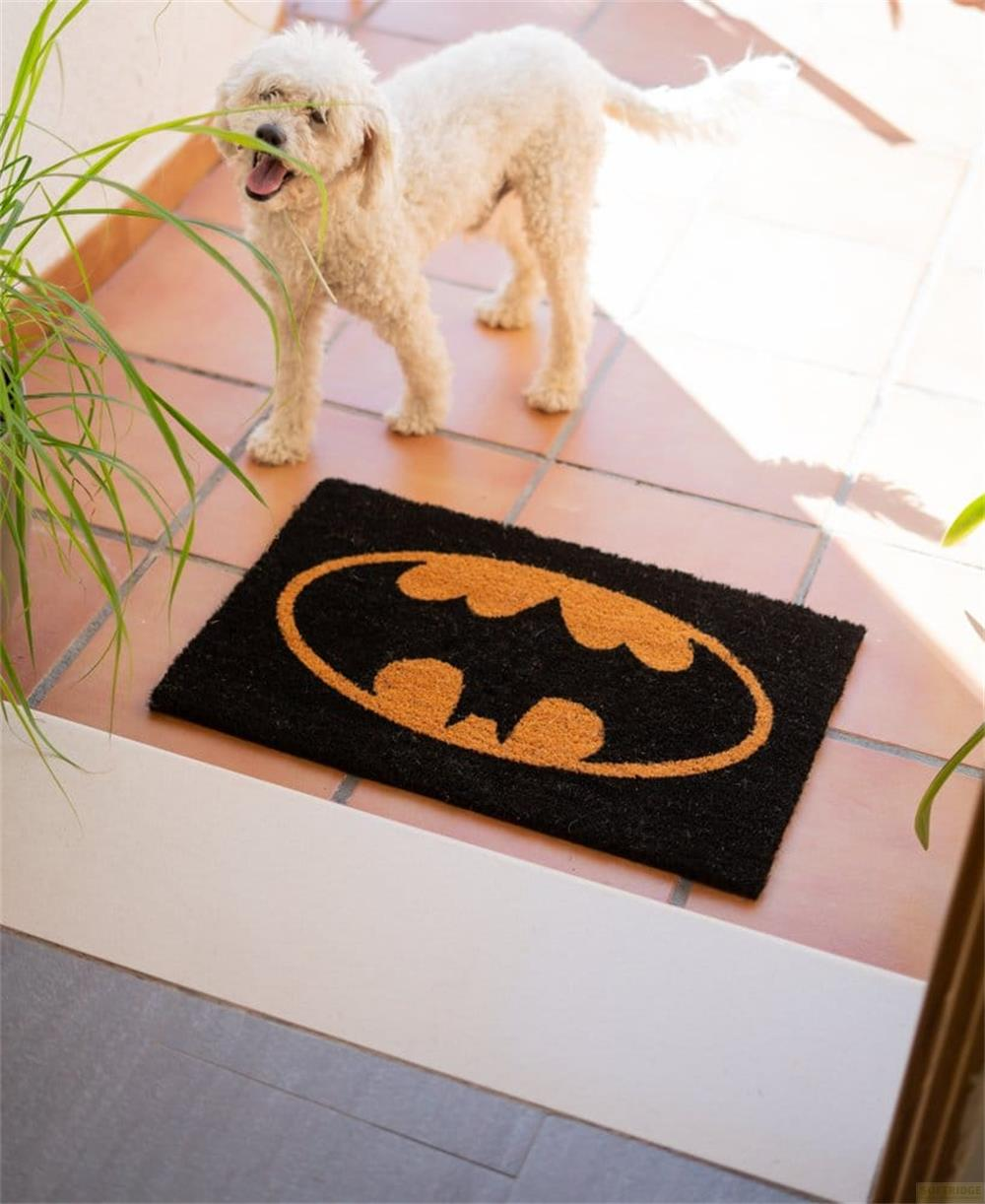 Ein weißer Pudel steht neben einer schwarzen Fußmatte mit dem Batman-Logo.