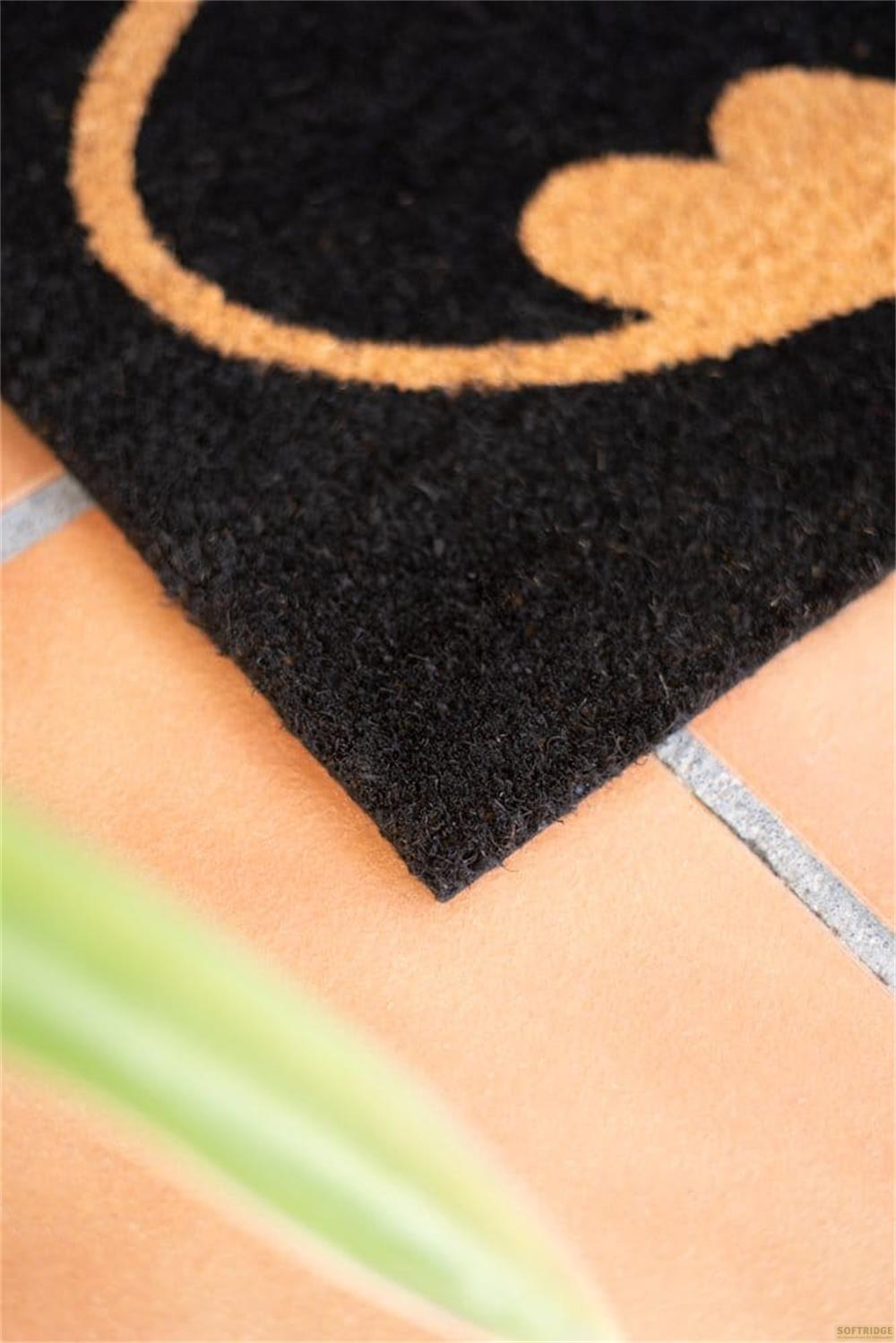 Schwarzer Teppich mit orangefarbenem Muster auf Kacheln. Grüne Pflanze im Vordergrund.