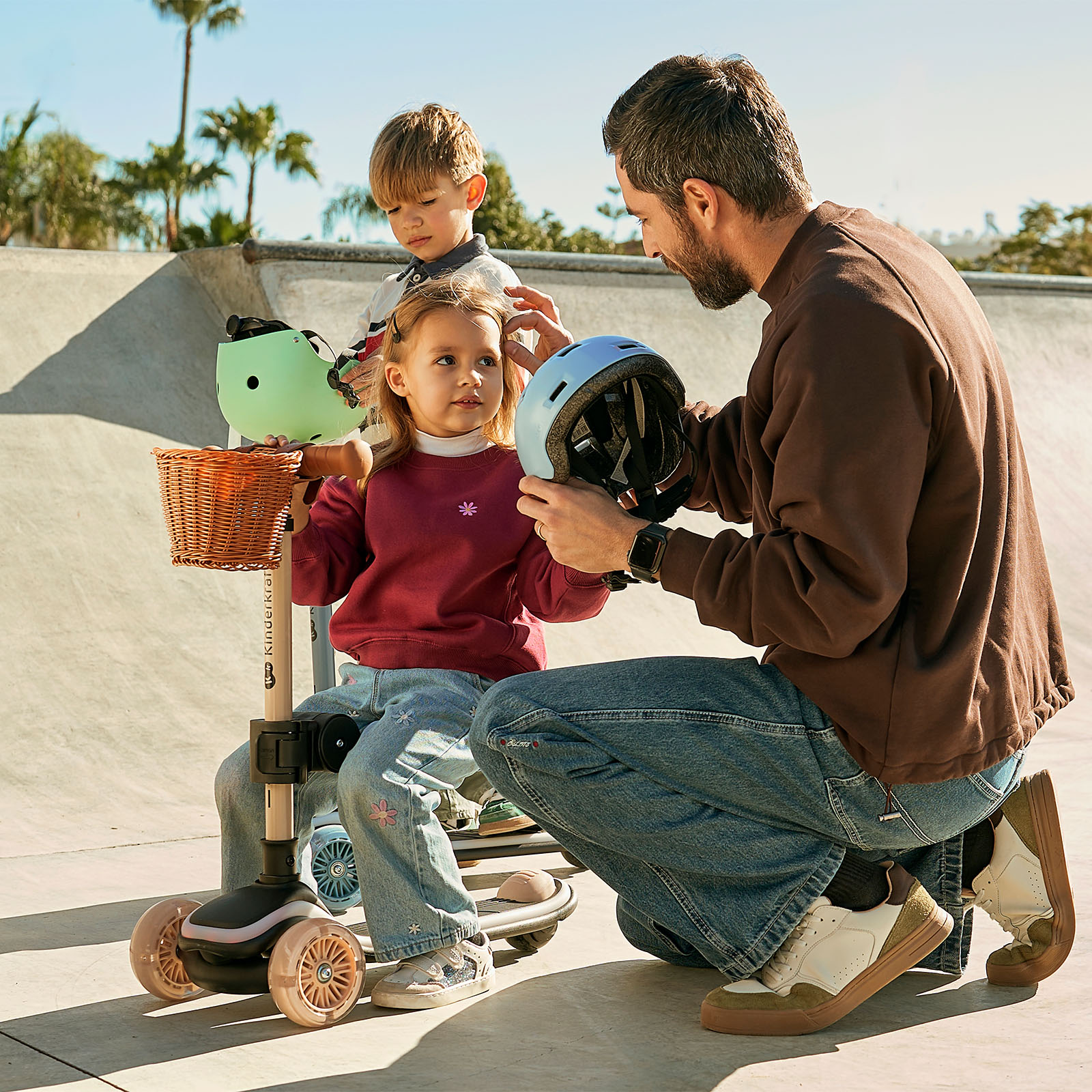 Ein Mann hilft zwei Kindern, Helme aufzusetzen, auf einem Skateboard-Übungsgelände.