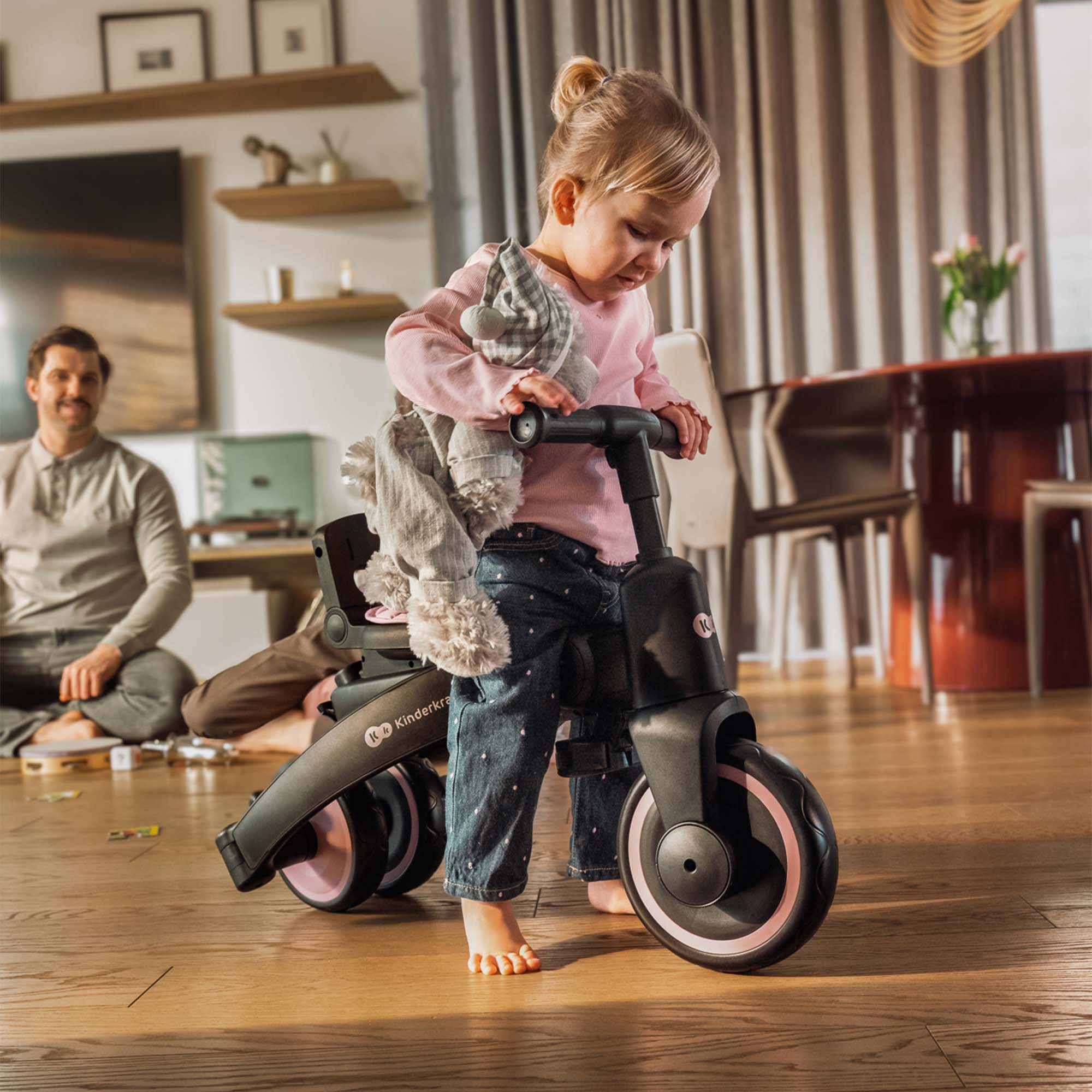 Una niña con jeans monta un patinete triciclo con un peluche.