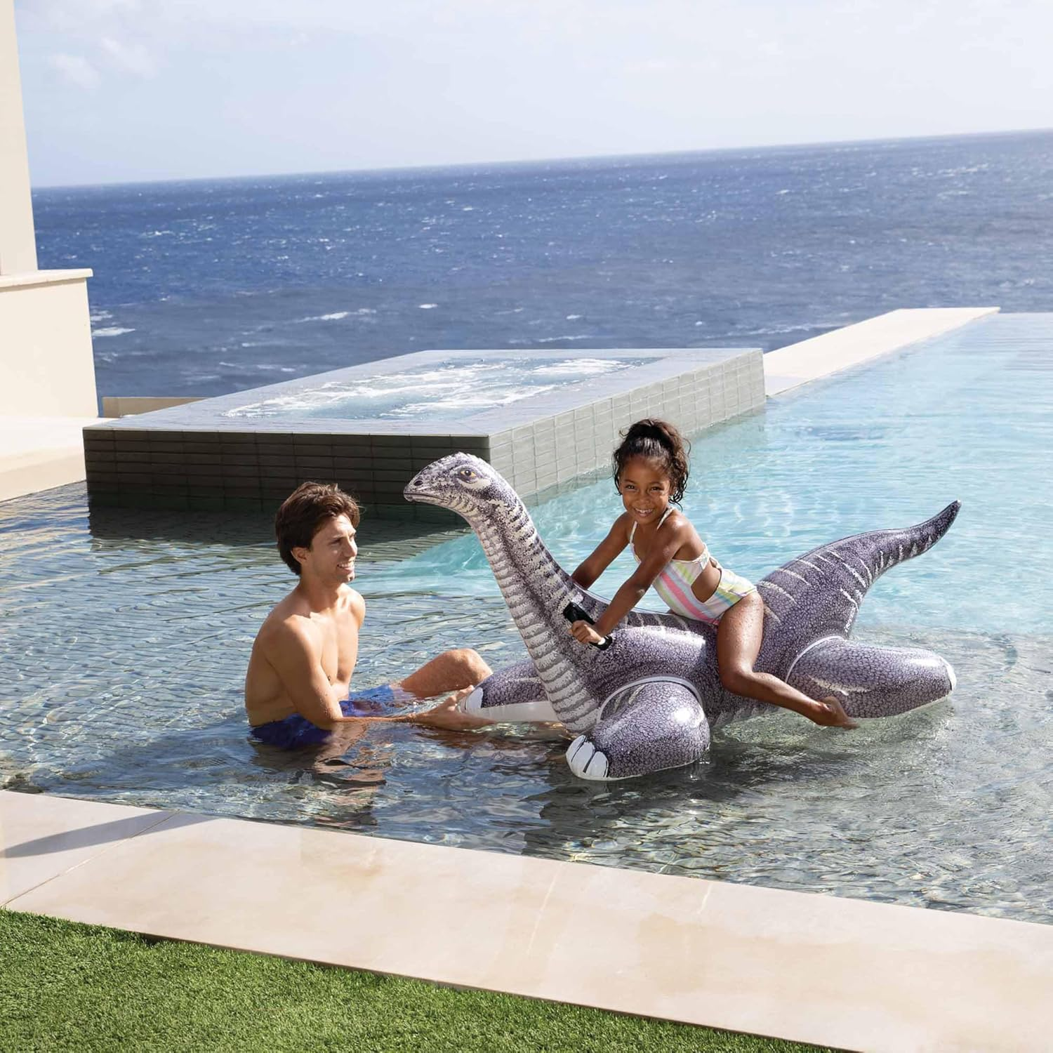 Ein Mann und ein Mädchen reiten auf einem aufblasbaren Dinosaurier in einem Pool am Meer.