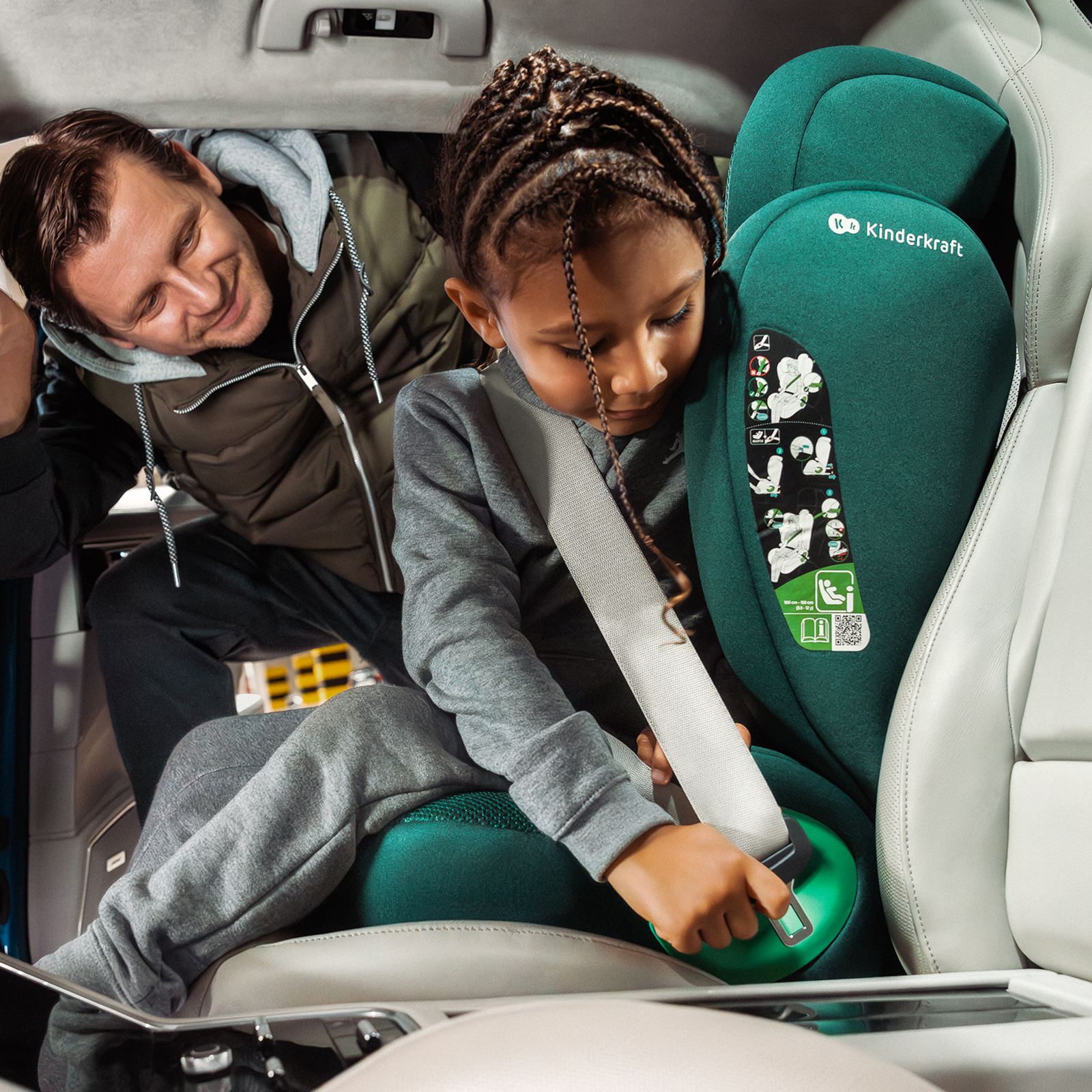 Mann hilft Kind im Autositz sich anzuschnallen. Dunkelgrüner Sitz.
