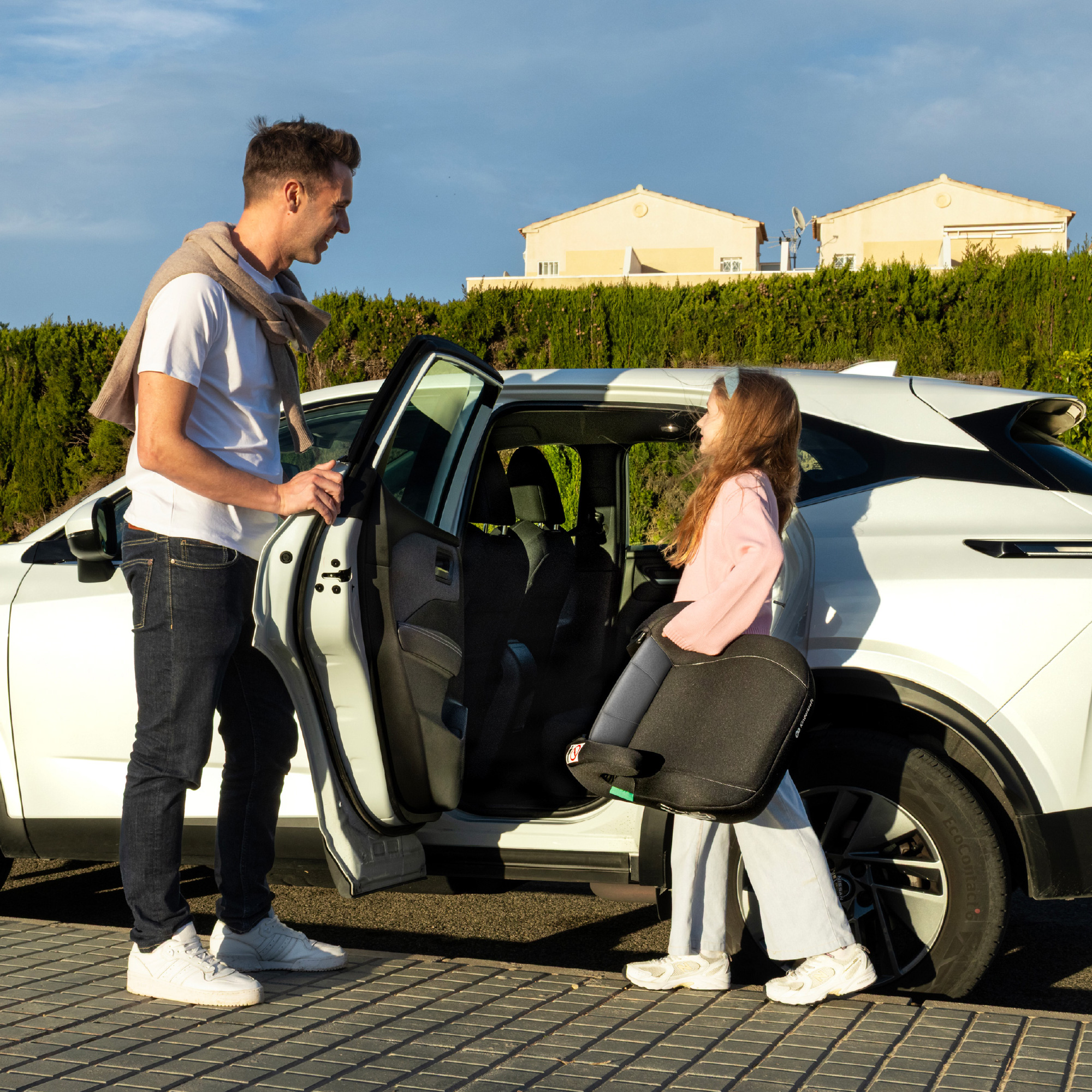 Mann öffnet Autotür für Mädchen mit Kindersitz. Sonnenschein, Häuser im Hintergrund.
