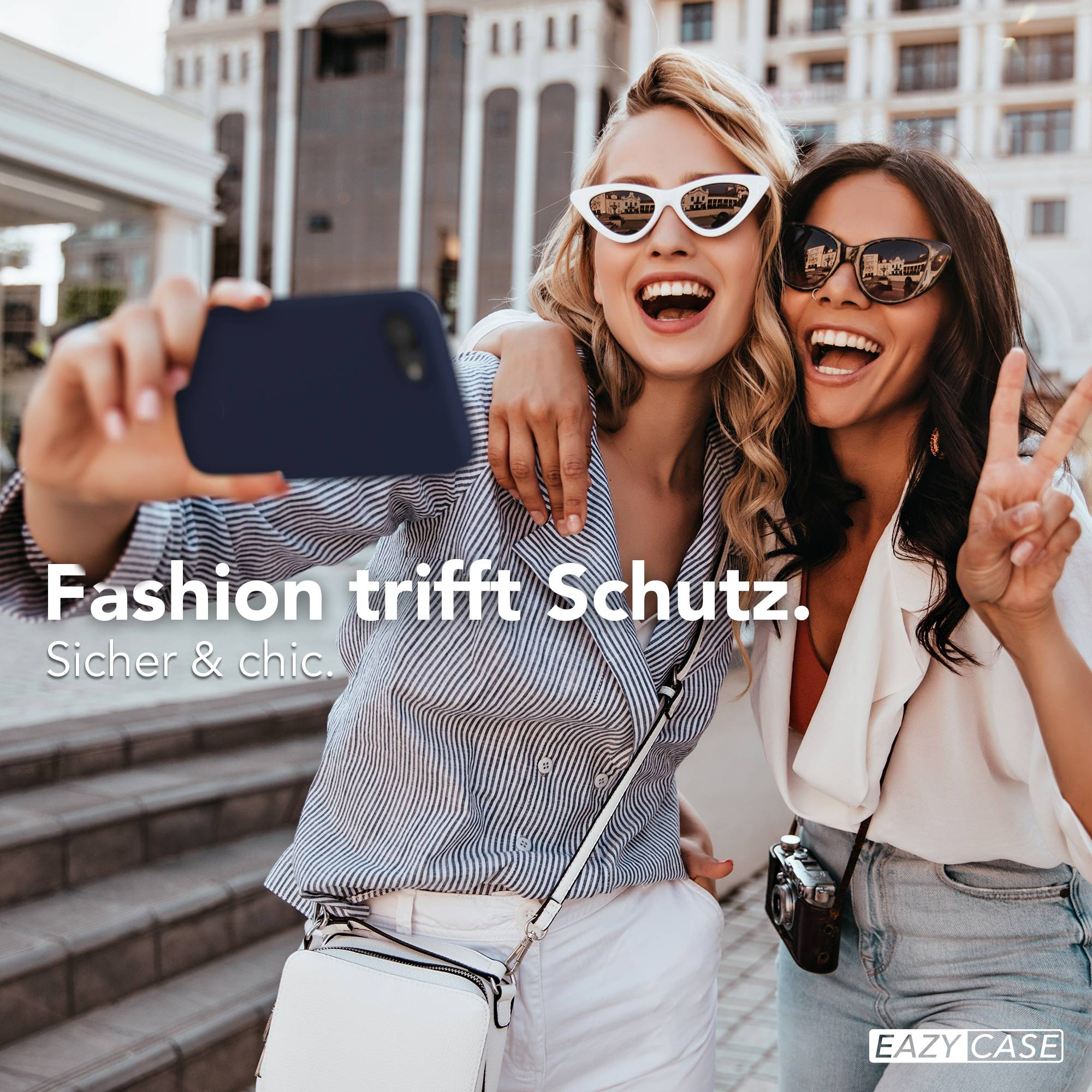 Zwei lachende Frauen machen ein Selfie vor einem Gebäude. Sie tragen Sonnenbrillen.