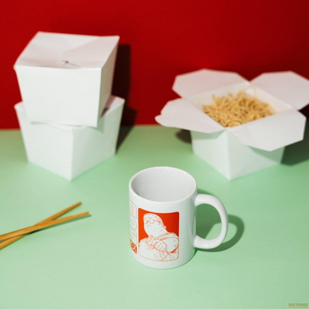 Taza blanca con gráfico naranja, palillos y cajas de fideos sobre superficie verde claro.