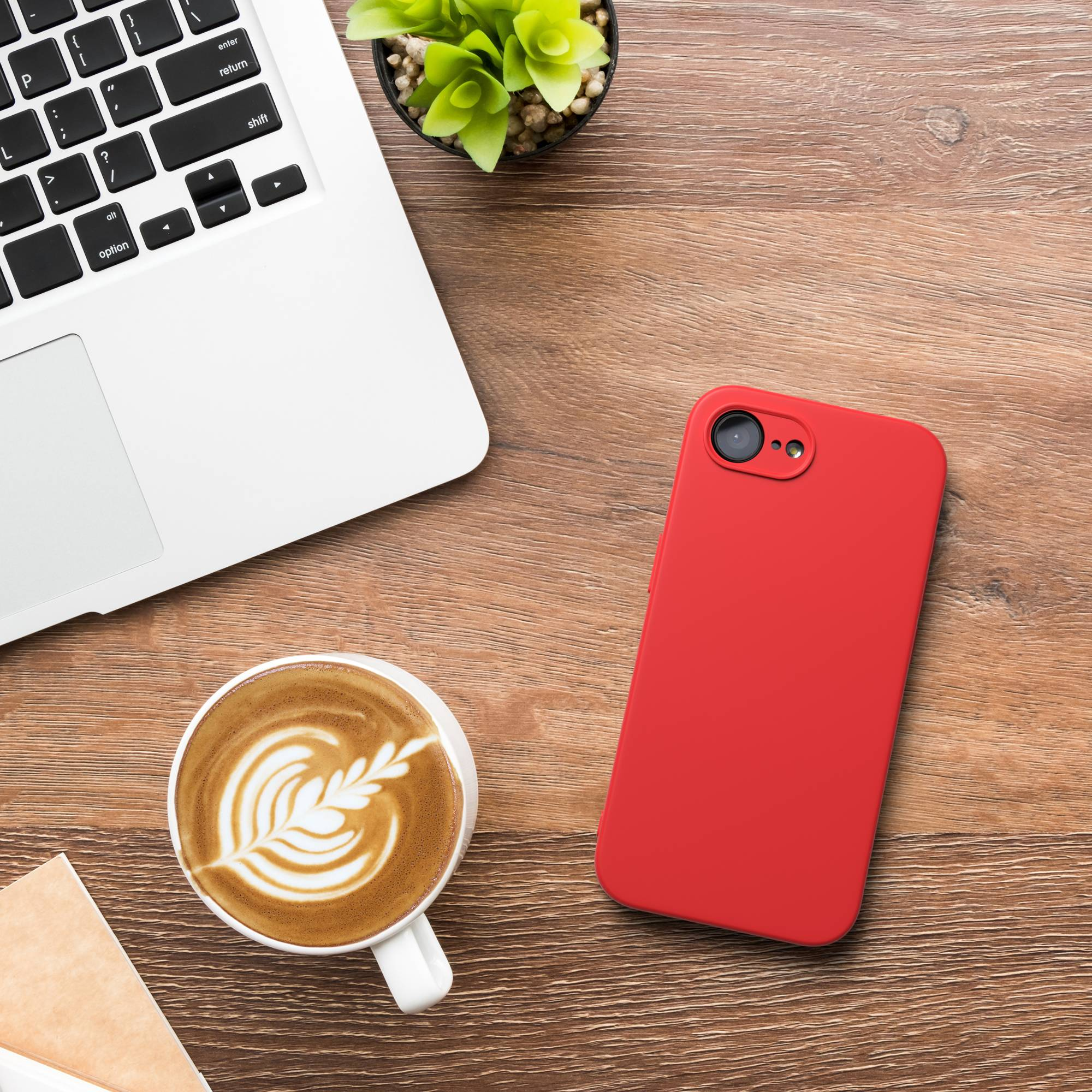 Ein Laptop, eine rote Handyhülle, eine Kaffeetasse und eine Pflanze auf einem Holztisch.