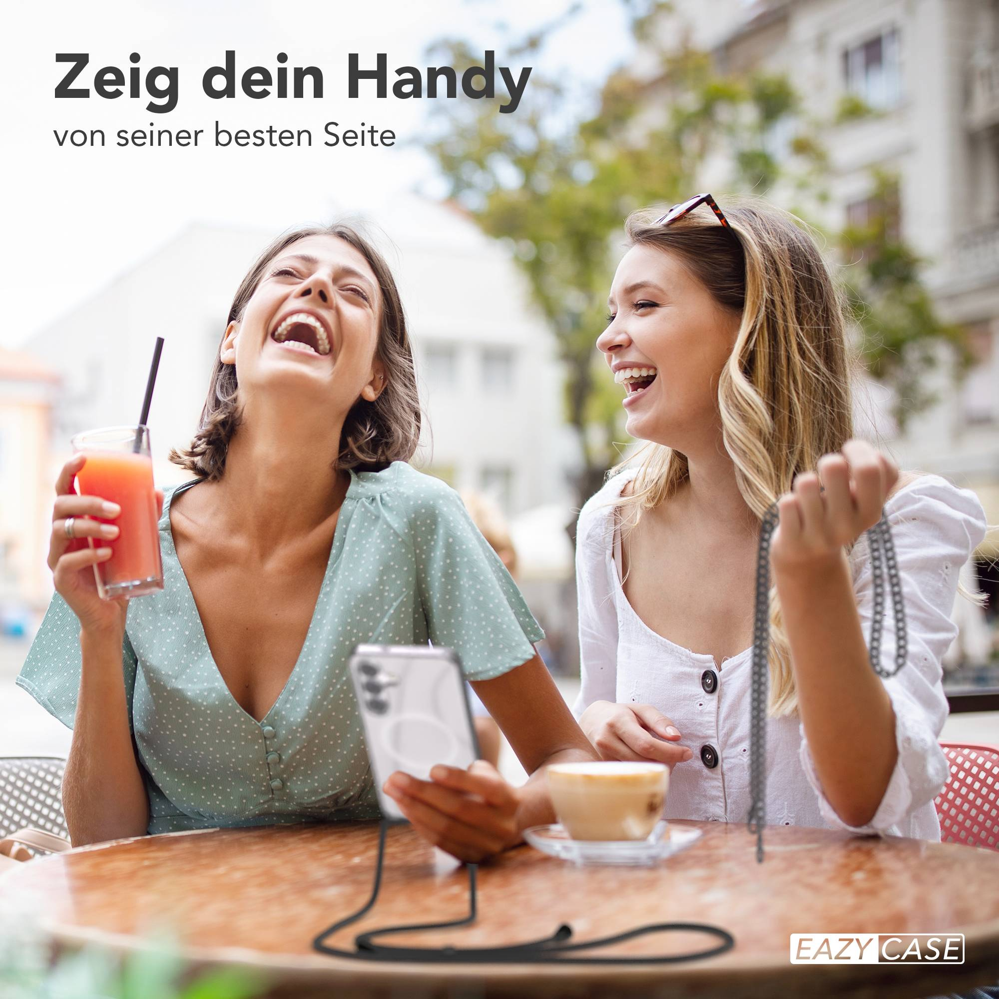 Zwei lachende Frauen sitzen an einem Tisch im Freien. Eine hält ein Getränk, die andere eine Kette.