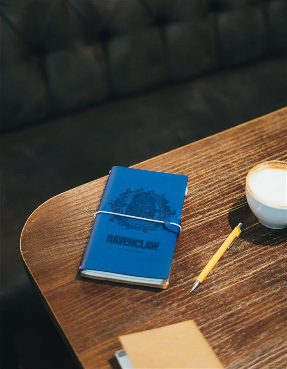 Ein blaues Ravenclaw-Notizbuch, ein gelber Stift und eine Kaffeetasse auf einem Holztisch.