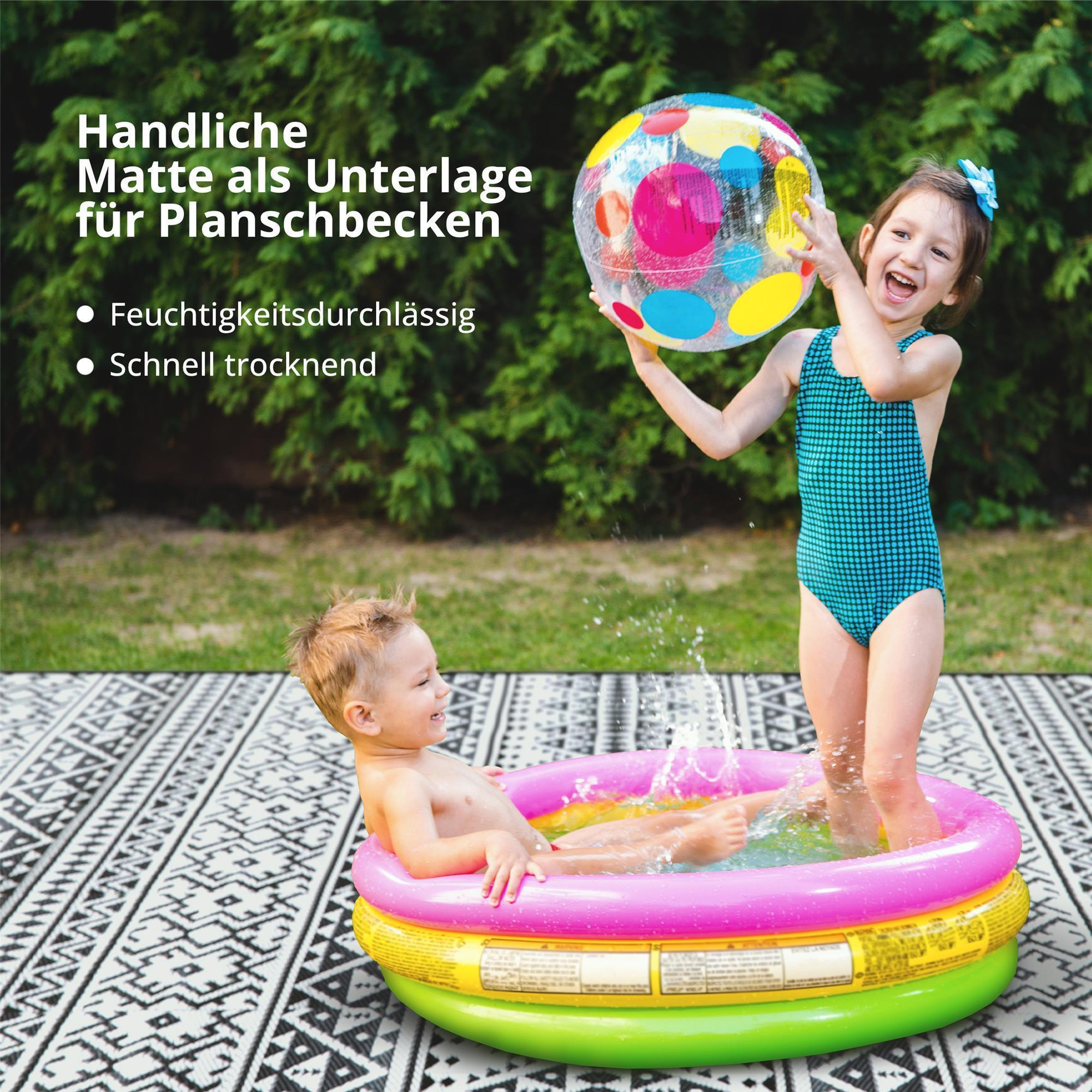 Zwei Kinder spielen in einem Planschbecken auf einer bunten Matte im Garten.