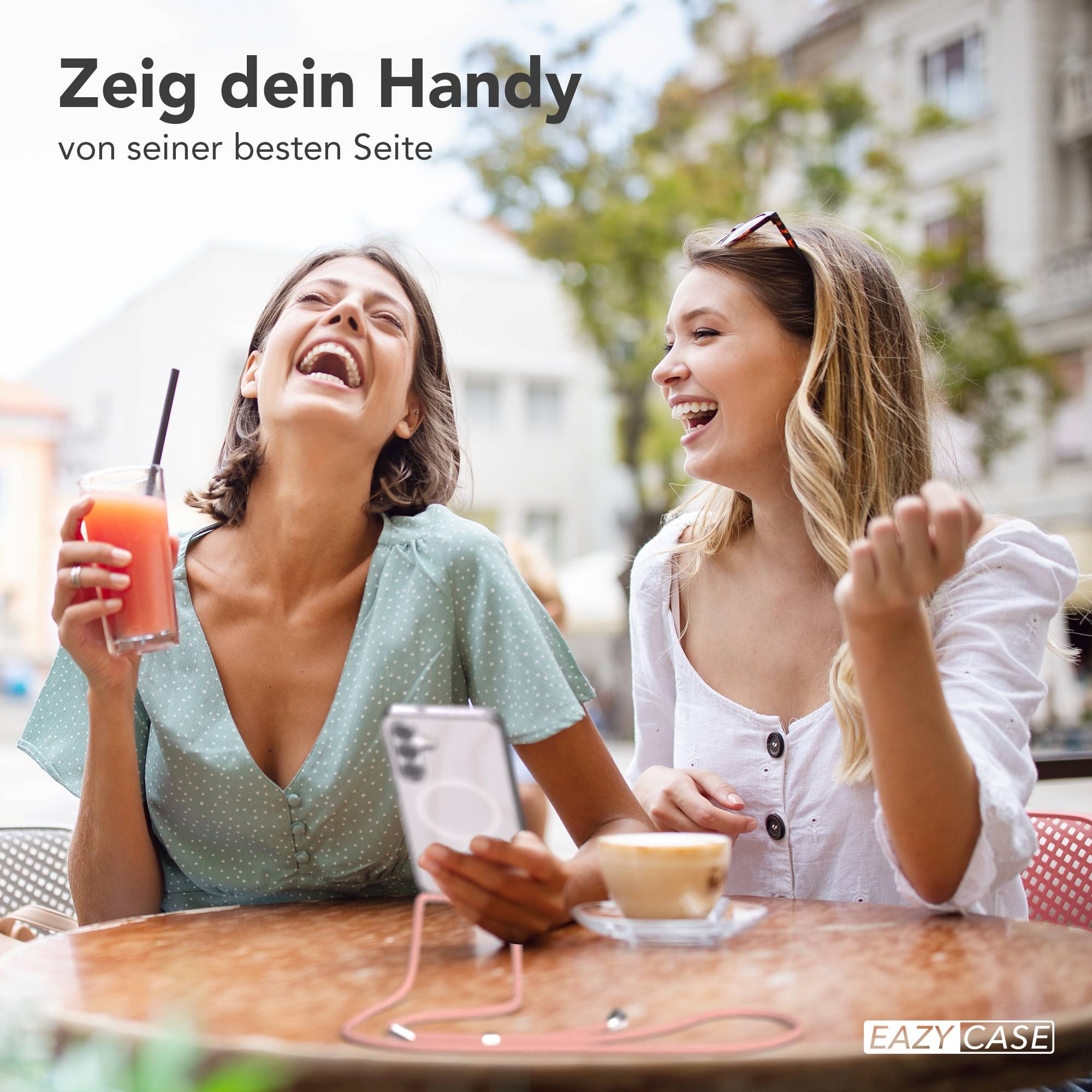 Zwei lachende Frauen sitzen an einem Tisch im Freien mit Getränken und einem Handy.
