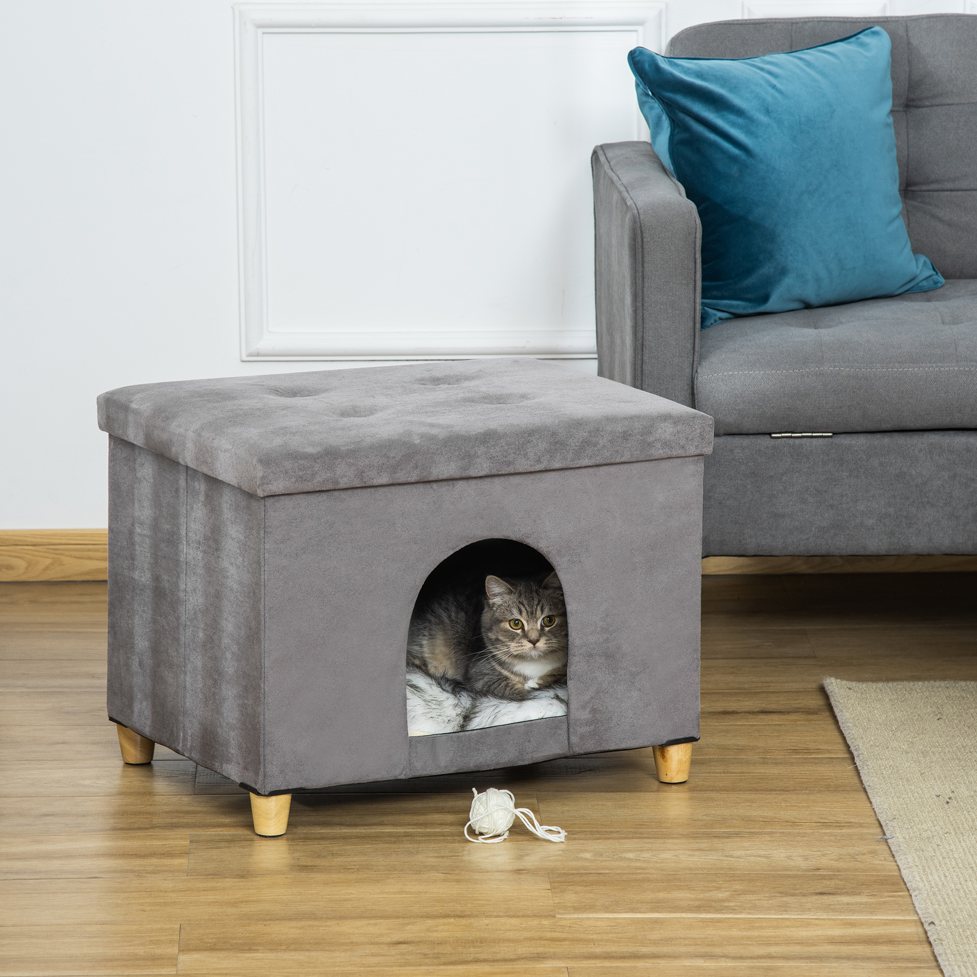 Una casa gris para gatos con un gatito dentro, junto a una bola de lana.