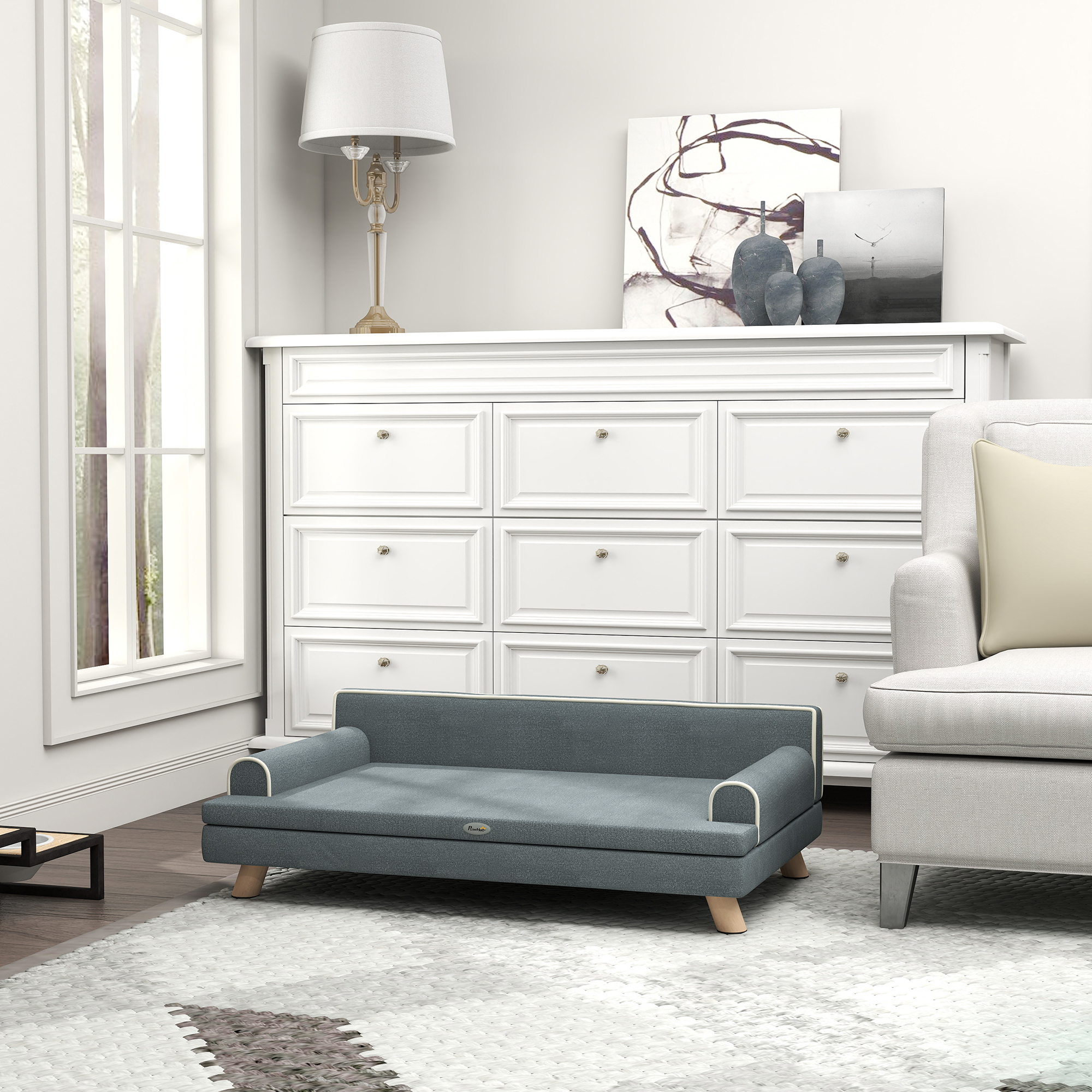 Una cama para perros gris se sienta frente a una cómoda blanca con arte y un sillón.