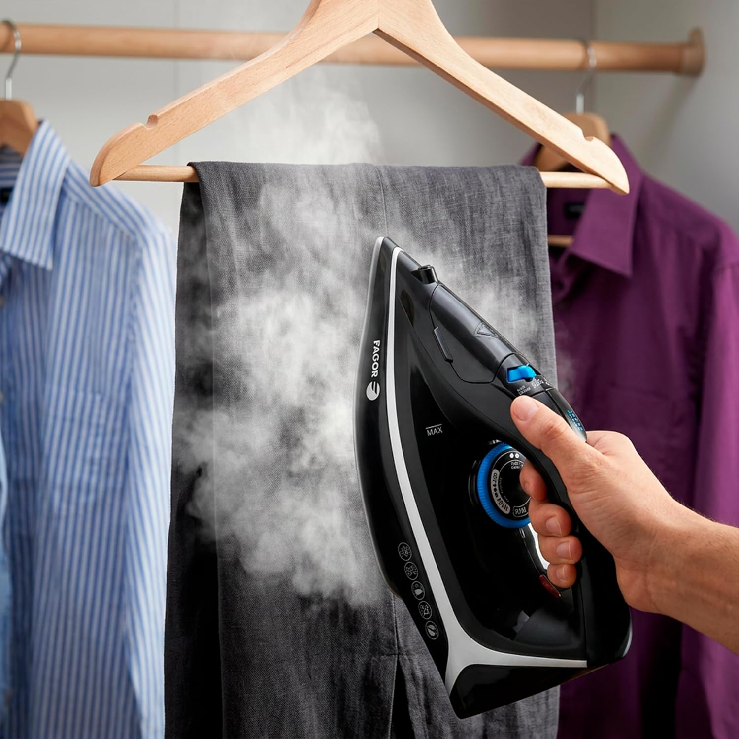 Mano plancha pantalones grises con vapor; camisa azul rayada y morada cerca.