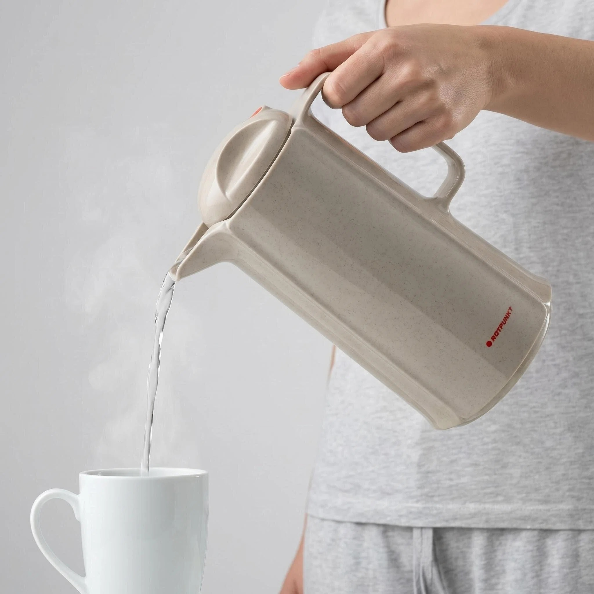 Eine Hand gießt heißes Wasser aus einem beigen Krug in eine weiße Tasse.
