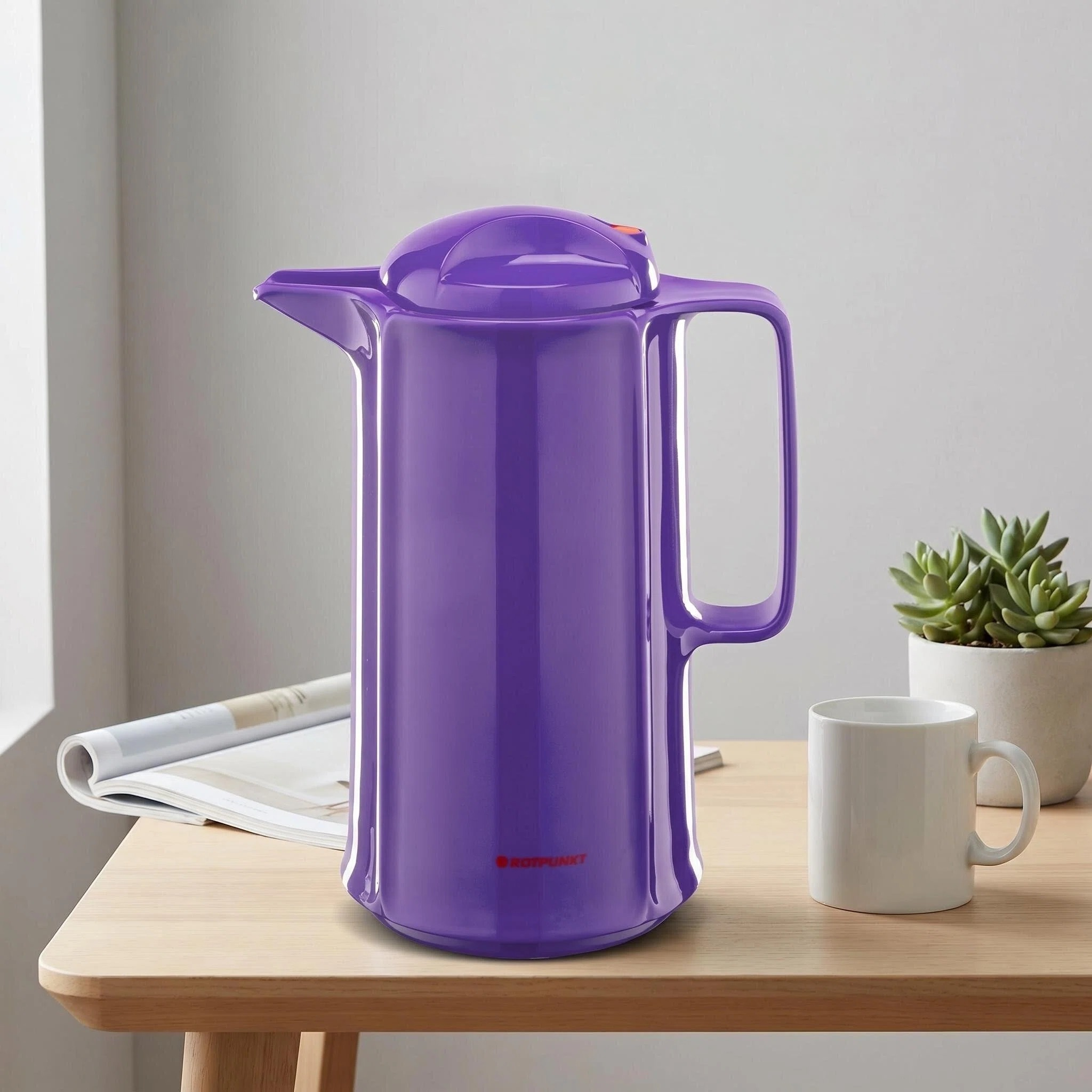 Eine lila Thermoskanne steht neben einer weißen Tasse und einem Magazin auf einem Holztisch.