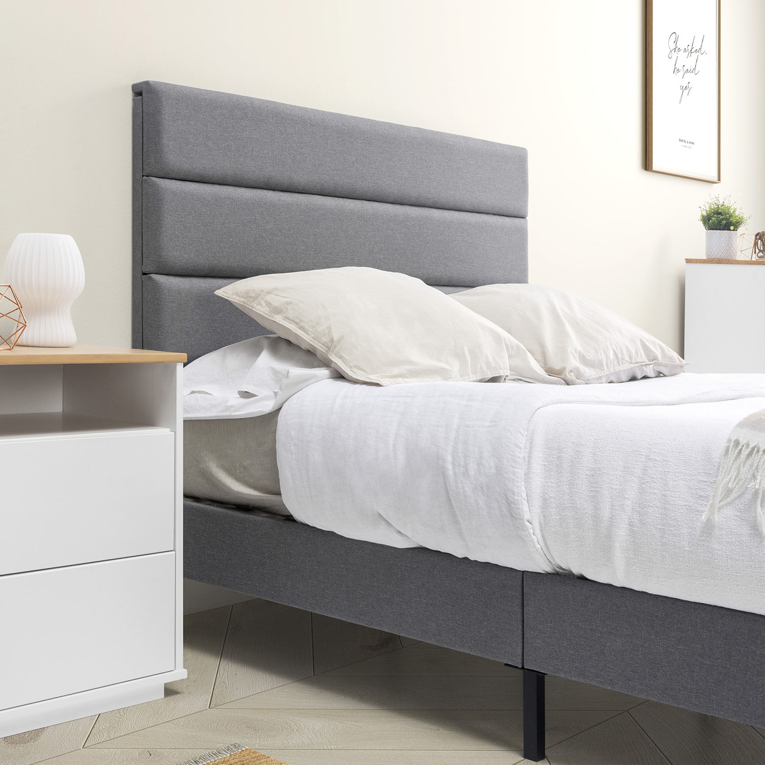 Una cama tapizada gris con sábanas y almohadas blancas, junto a una mesita de noche blanca.
