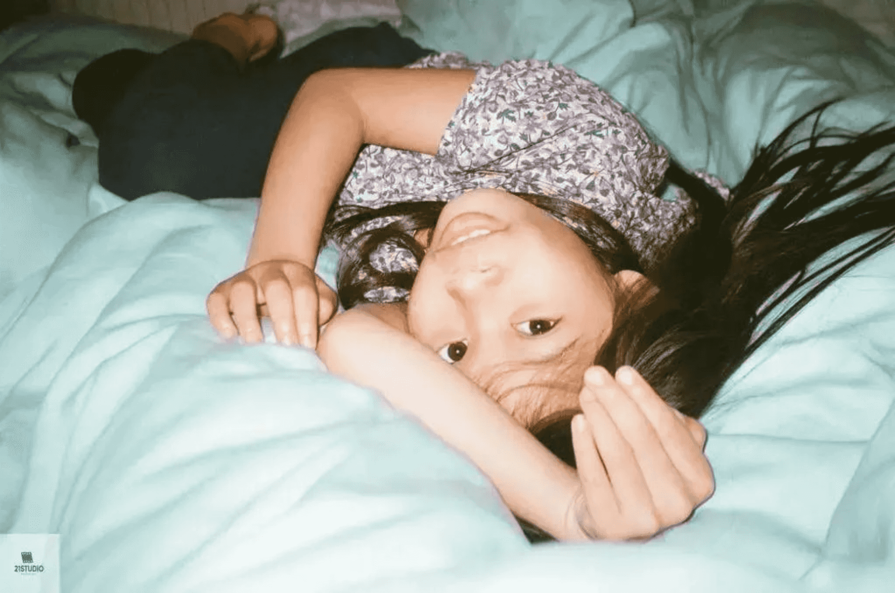 Una niña joven yace boca arriba en una cama azul claro, mirando a la cámara.