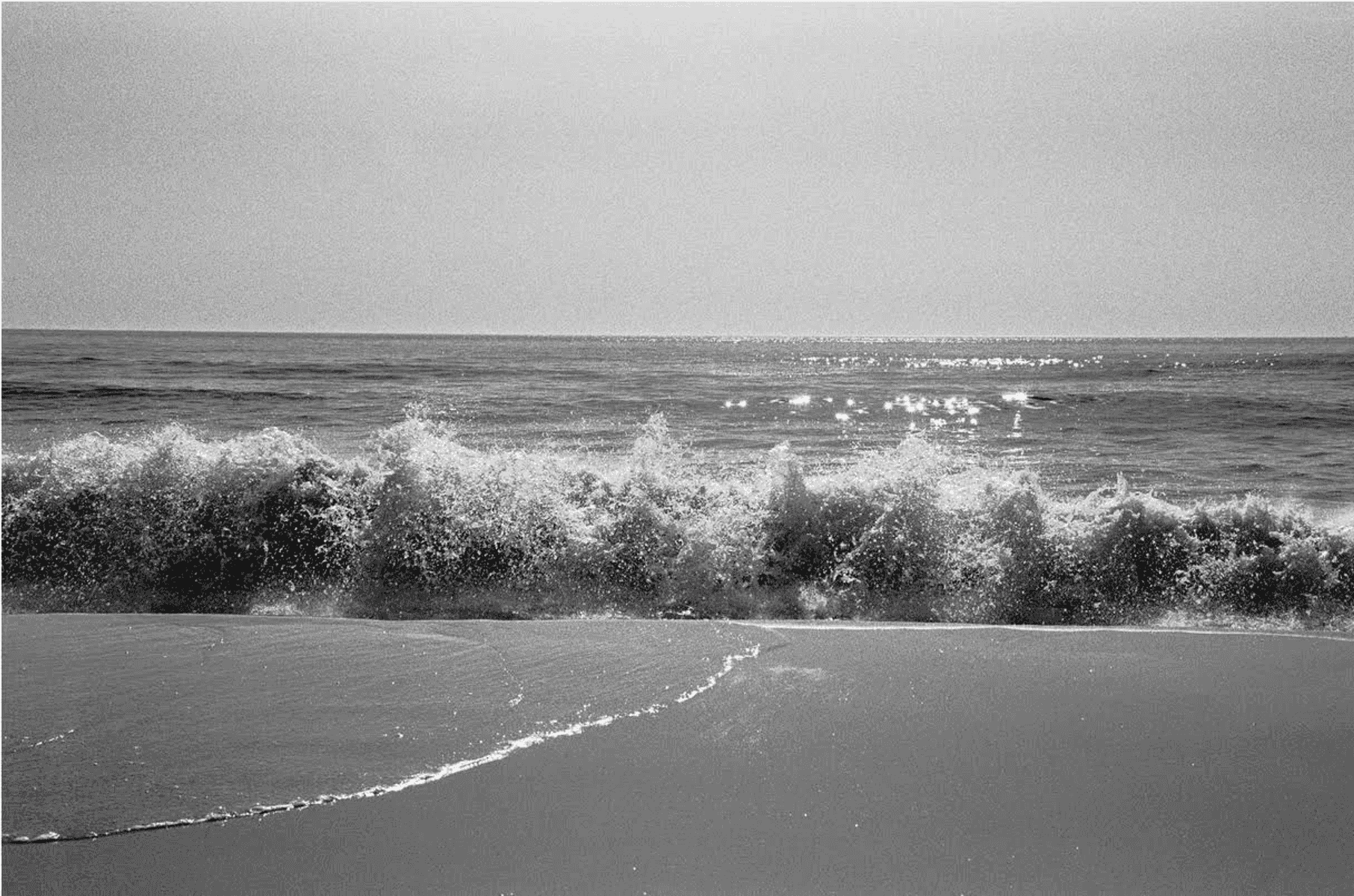 Eine brechende Welle am Strand mit Gischt und glitzerndem Wasser im Hintergrund.