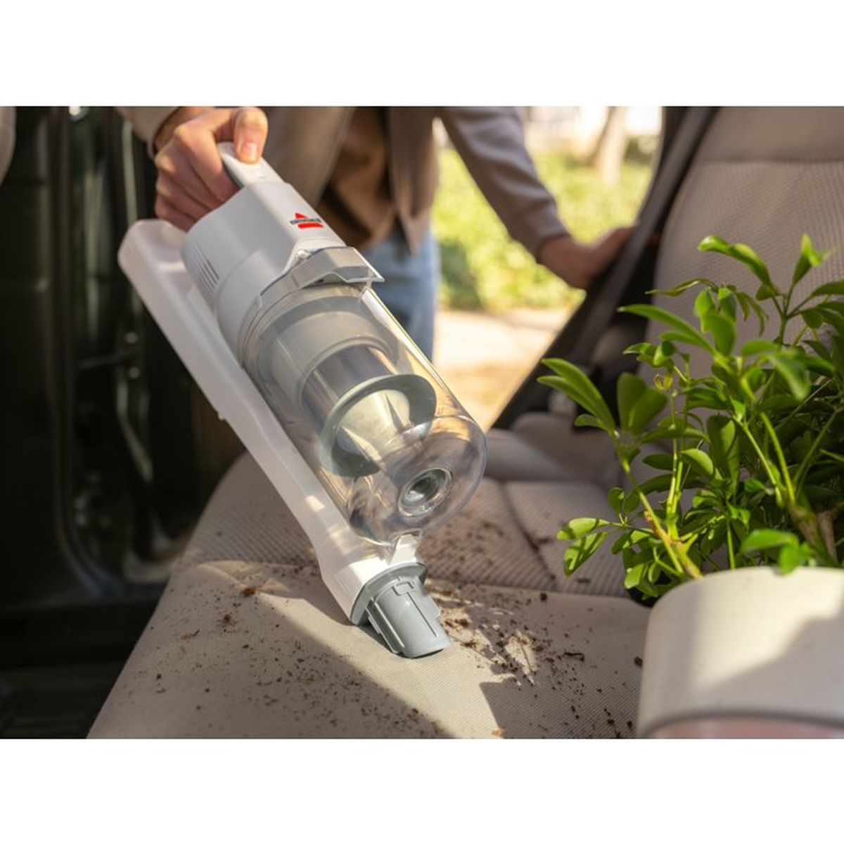 A hand holds a white handheld vacuum cleaning a car seat with dirt.