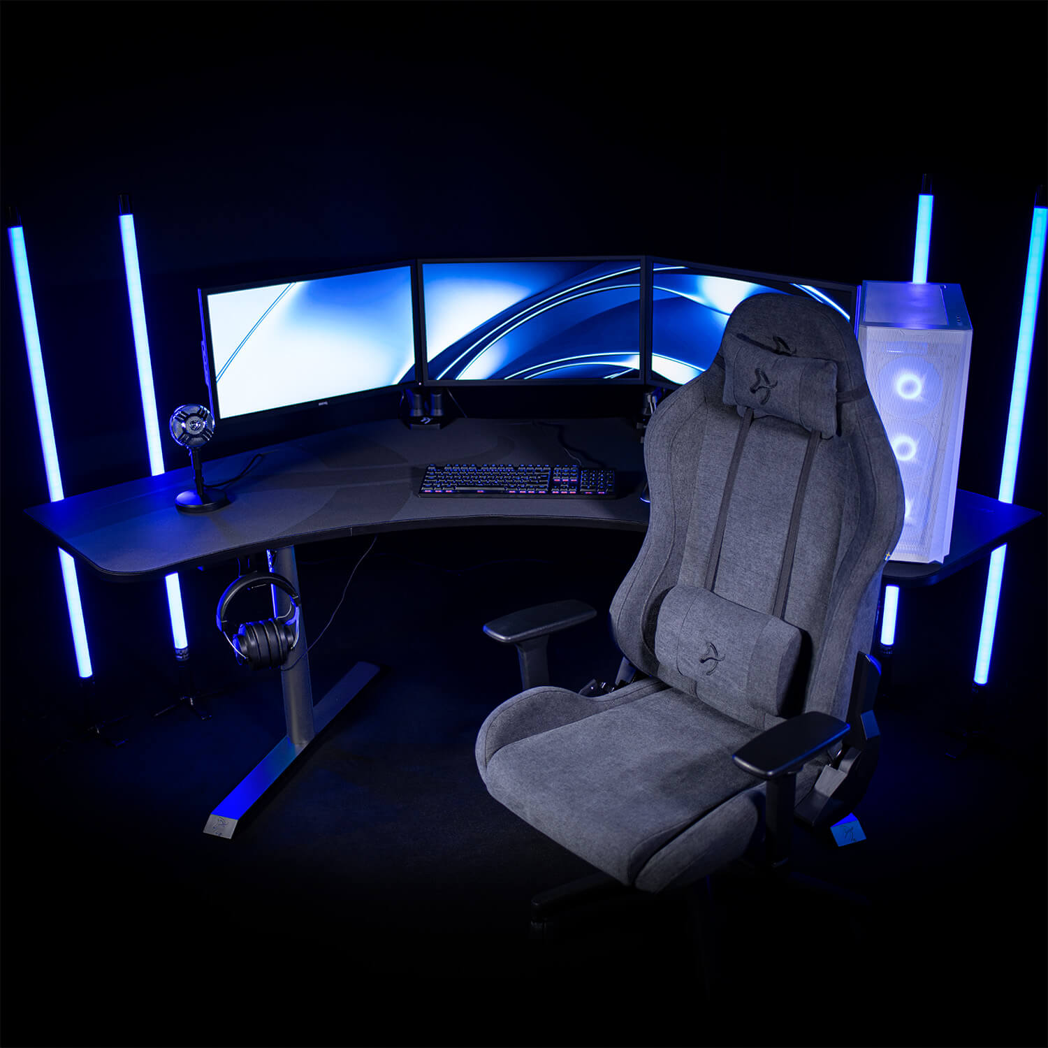 Una sedia da gaming grigia siede di fronte a una scrivania con tre monitor, tastiera, mouse e microfono.