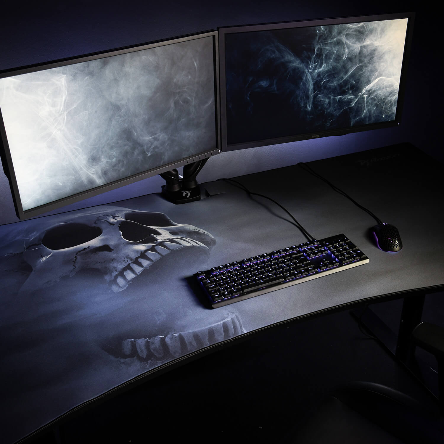 Due monitor sopra una tastiera, un mouse e un tappetino da scrivania con stampa di teschio.
