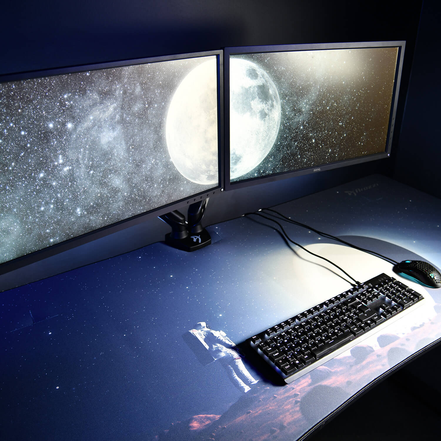 Doppi monitor mostrano luna e stelle. Tastiera e mouse su tappetino da scrivania a tema spaziale.