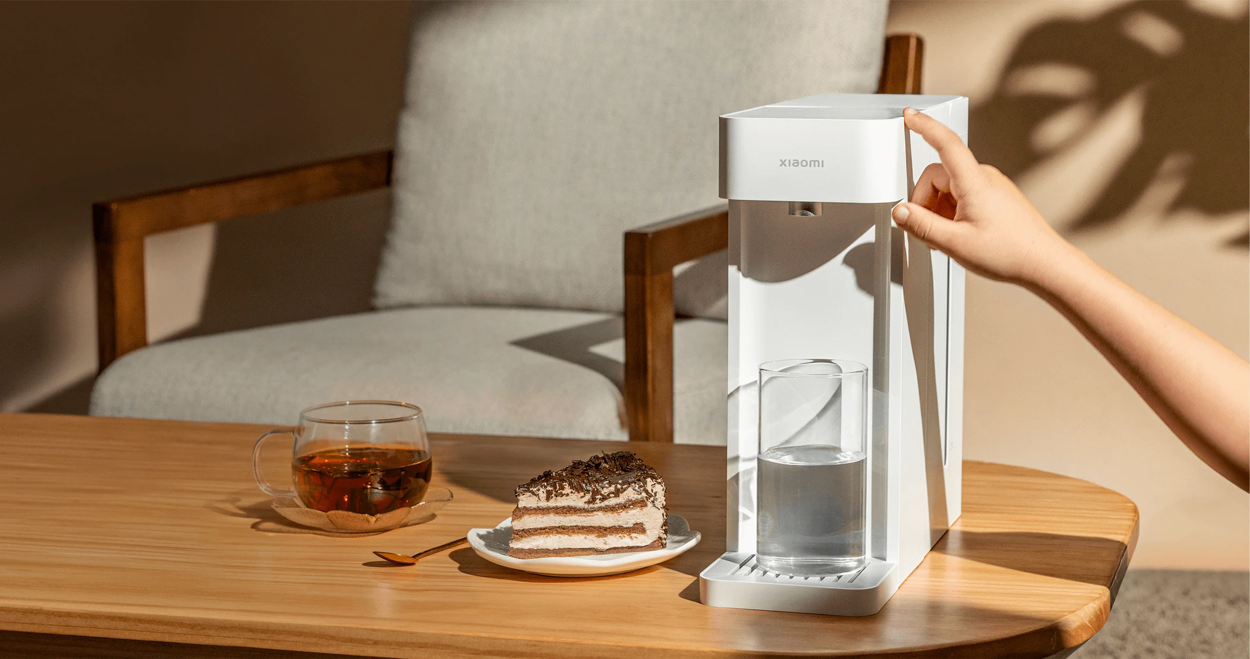 A finger presses a white water dispenser next to cake and tea.
