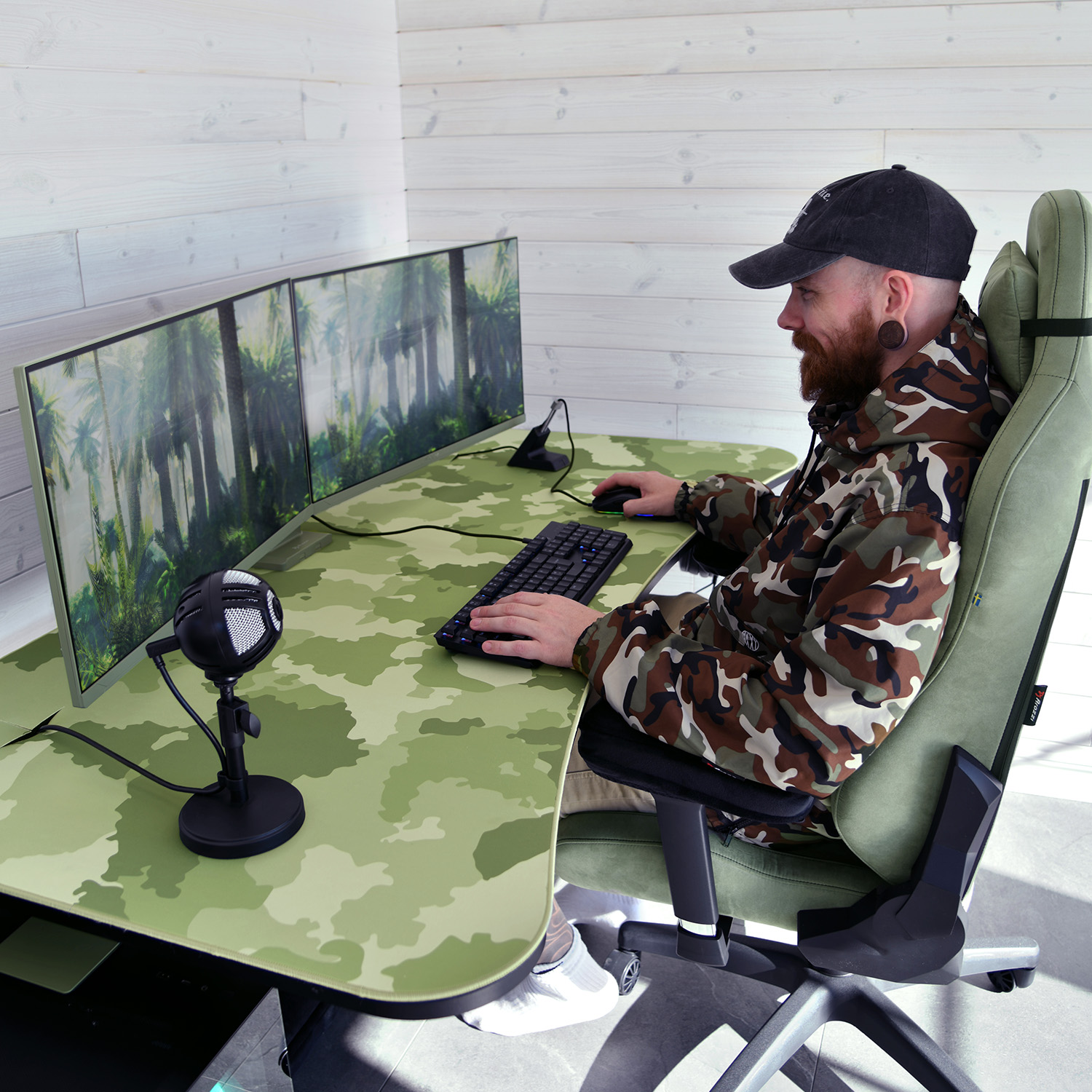Man in camouflage jacket sits at a desk with camouflage pattern, playing computer games.