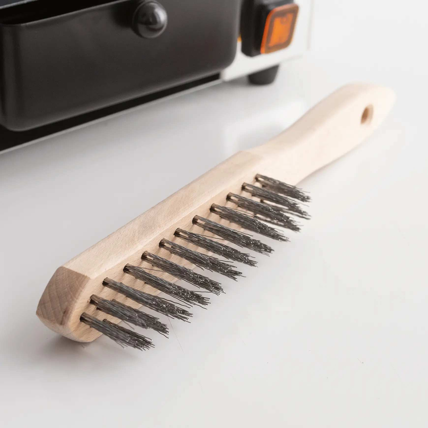 Drahtbürste mit Holzgriff liegt auf weißem Untergrund, daneben ein Teil eines Geräts.
