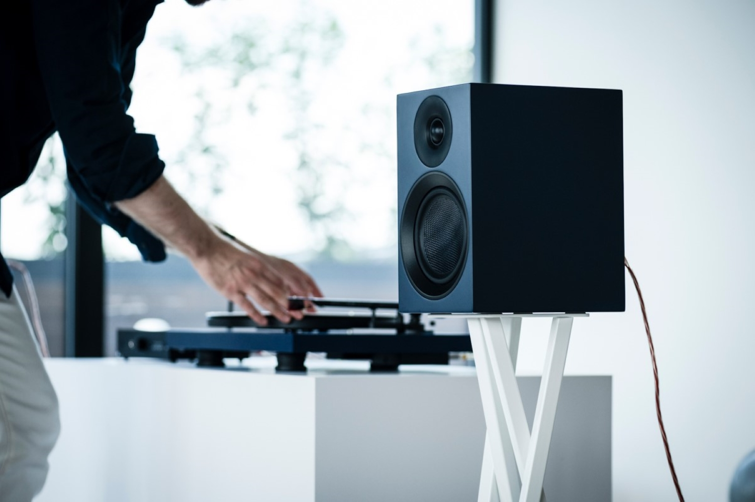 Mężczyzna obsługuje gramofon obok ciemnoniebieskiego głośnika na białym stojaku.