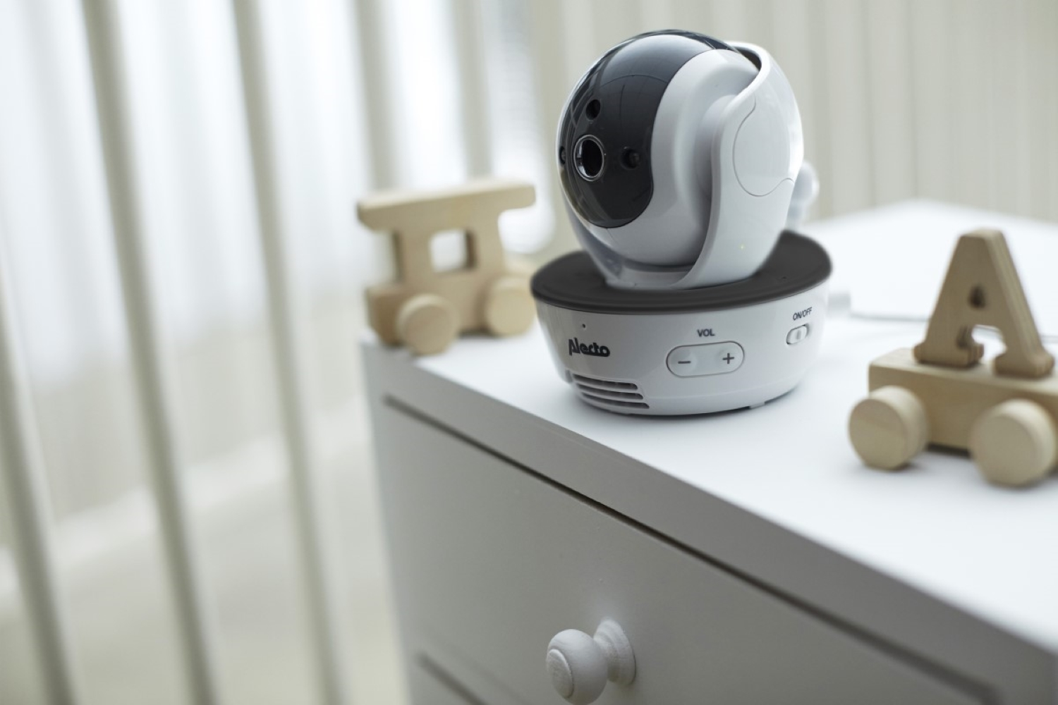 Biały monitor dla dziecka z czarną kamerą na białej komodzie, obok drewnianych zabawek.