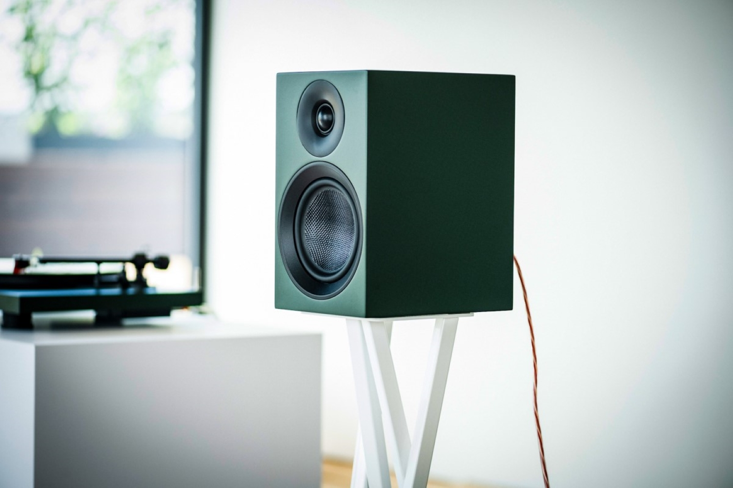 Ciemnozielony głośnik na białym stojaku obok gramofonu.