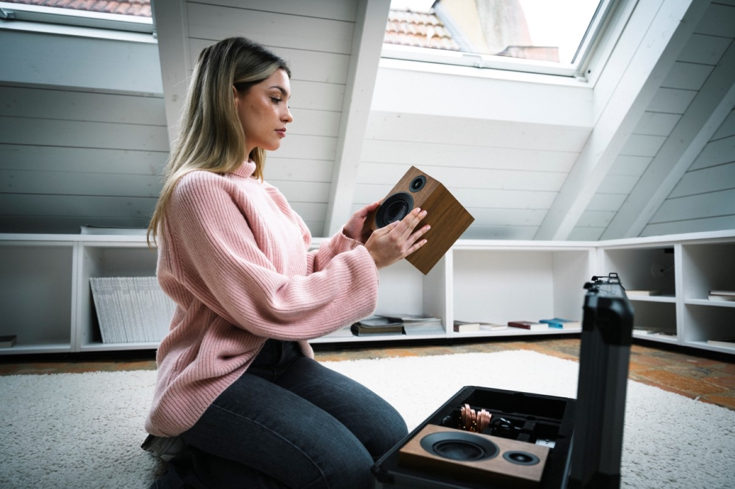 Kobieta w różowym swetrze trzyma drewniany głośnik na poddaszu.